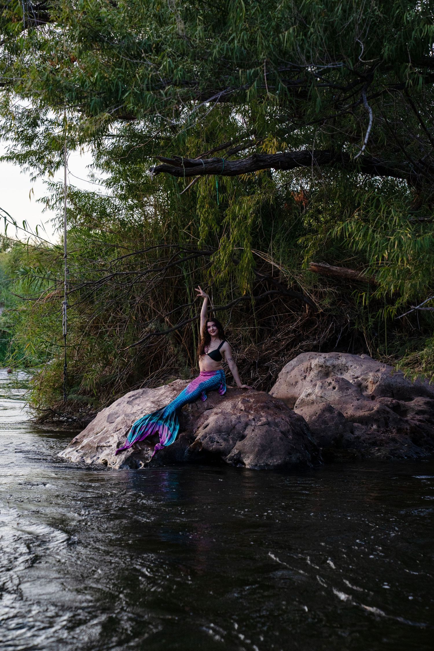 A woman in a mermaid costume poses dramatically on riverside rocks, raising her arm toward overhanging tree branches. Fantasy Photoshoot