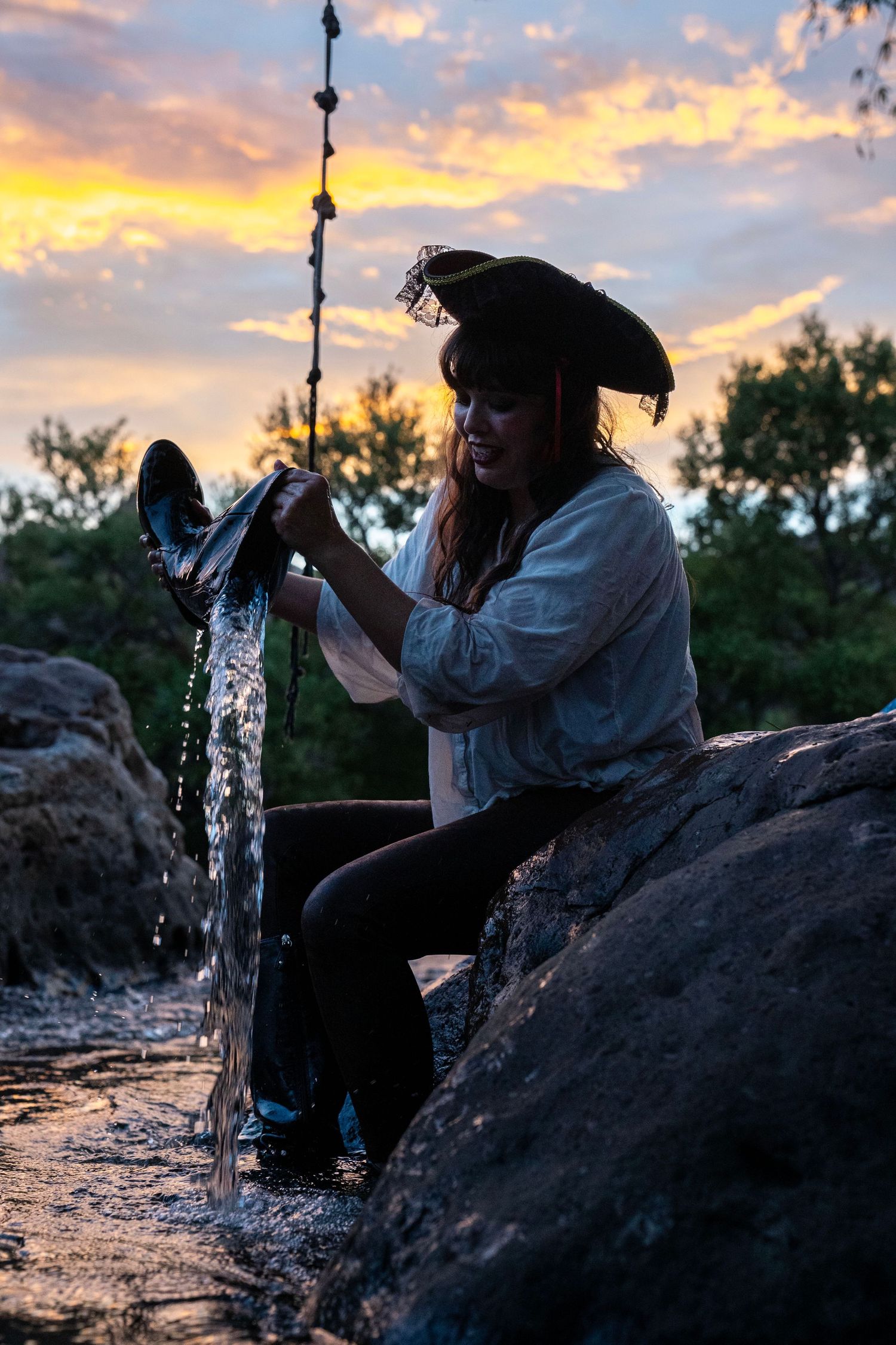 Pirate costume man empties water from boot on rocky riverbank at sunset. Mermaid photoshoot, fantasy photoshoot. Arizona photographer.