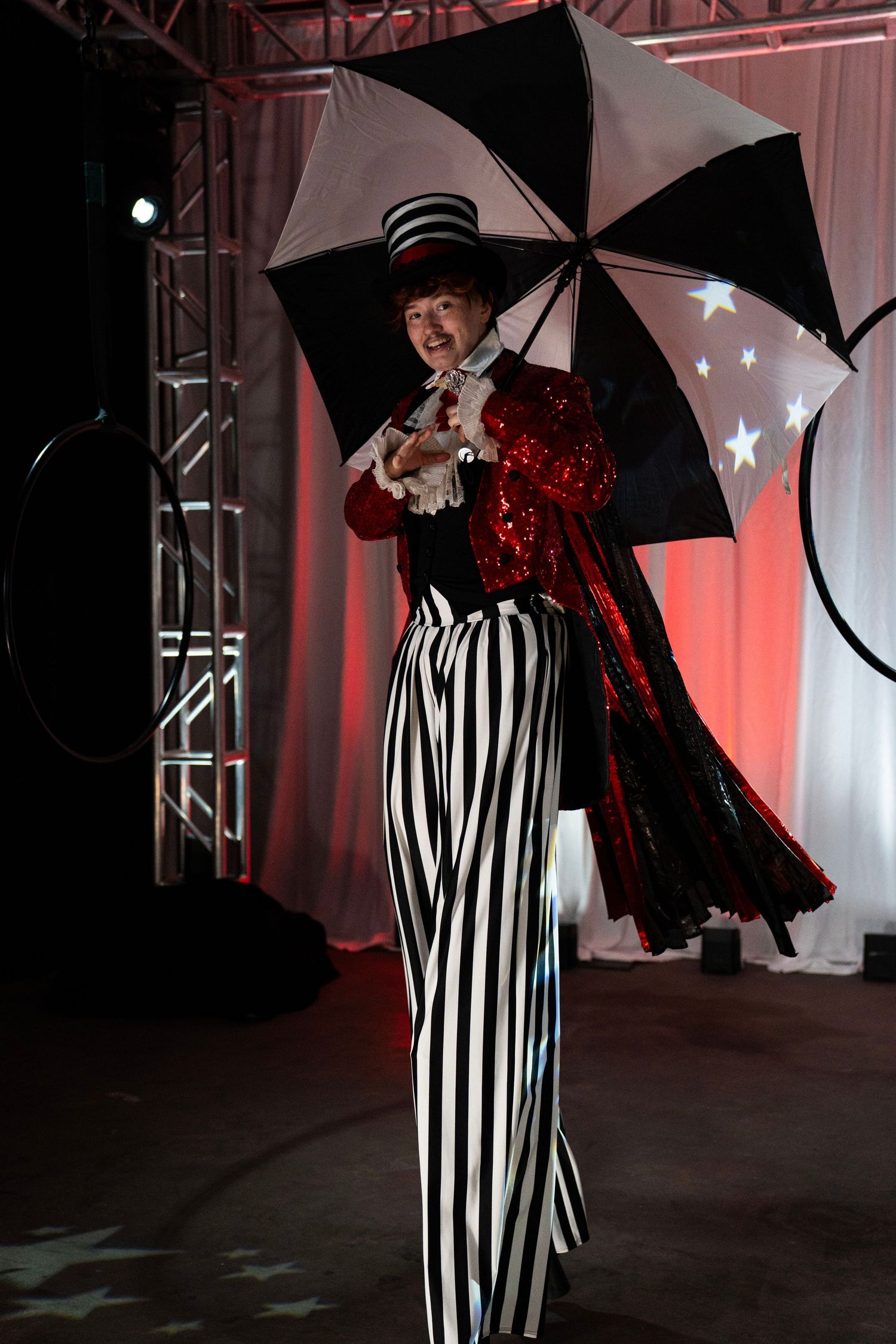 Female stilt walker in black and white striped costume holds umbrella at circus themed event.