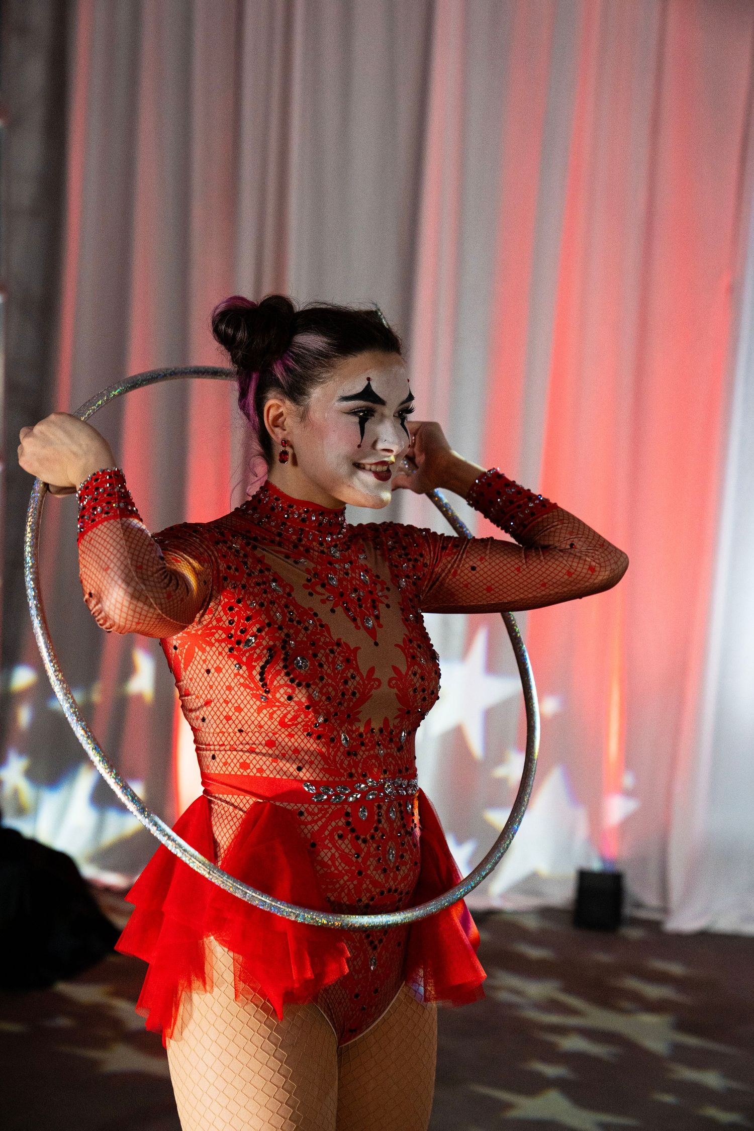 Circus performer with dramatic clown makeup holds silver hula hoop in red sequin costume on stage.