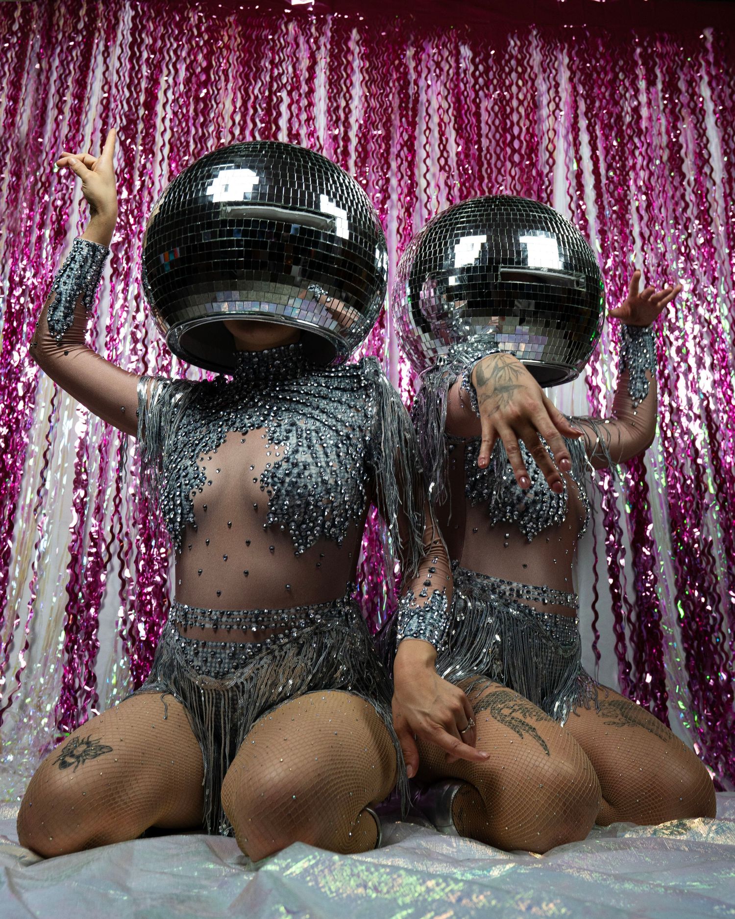 Two performers in sparkling silver fringe bodysuits with disco ball heads pose against pink tinsel.
