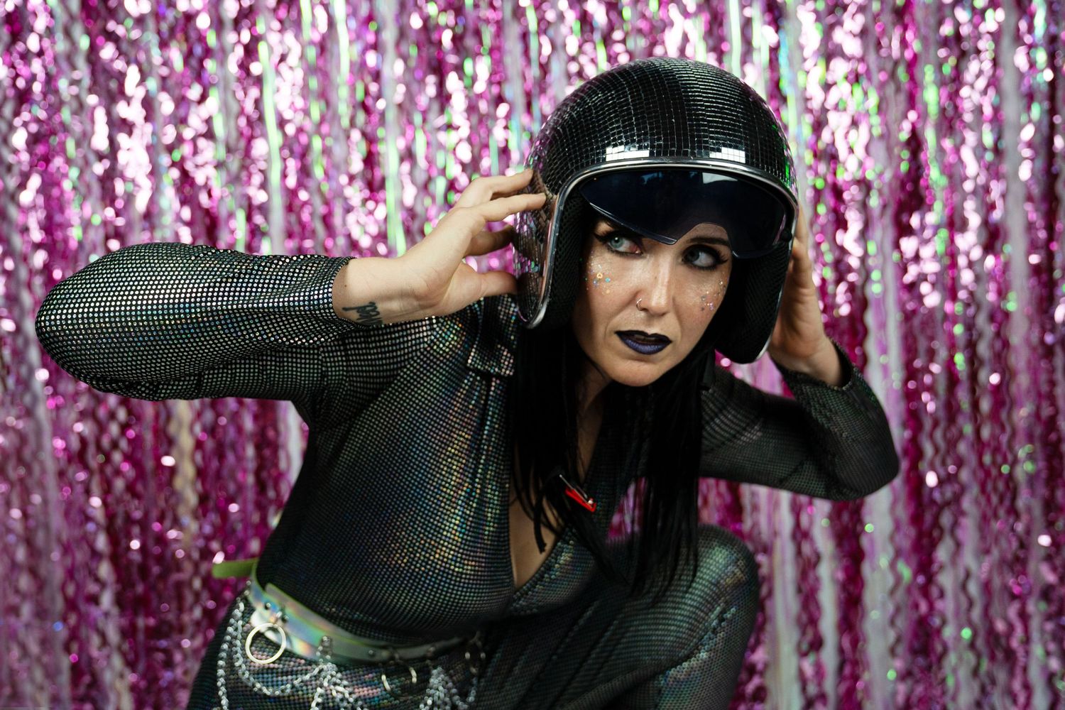A woman with dark makeup wears a black outfit and disco helmet against a pink sequin curtain.