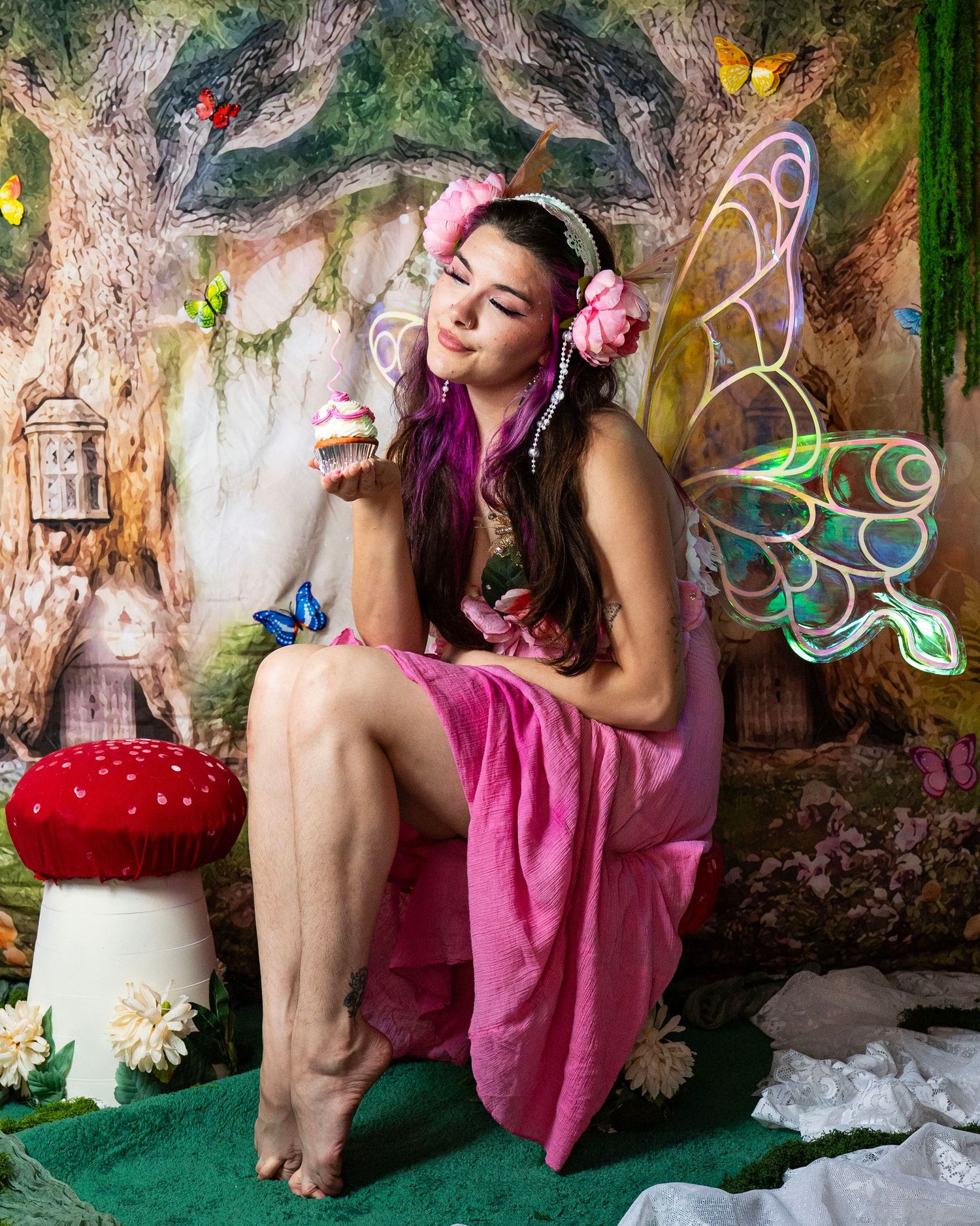 Barefoot fairy model in pink dress with holographic wings sits by red mushroom in magical forest backdrop. Fantasy Photoshoot