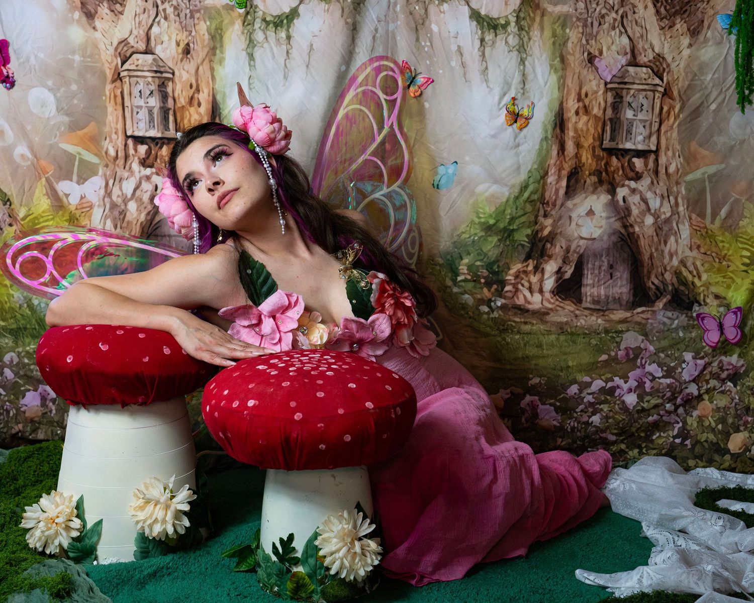 Woman dressed as fairy with pink wings and floral bodice reclines on giant red mushrooms in magical forest. Fantasy Photoshoot