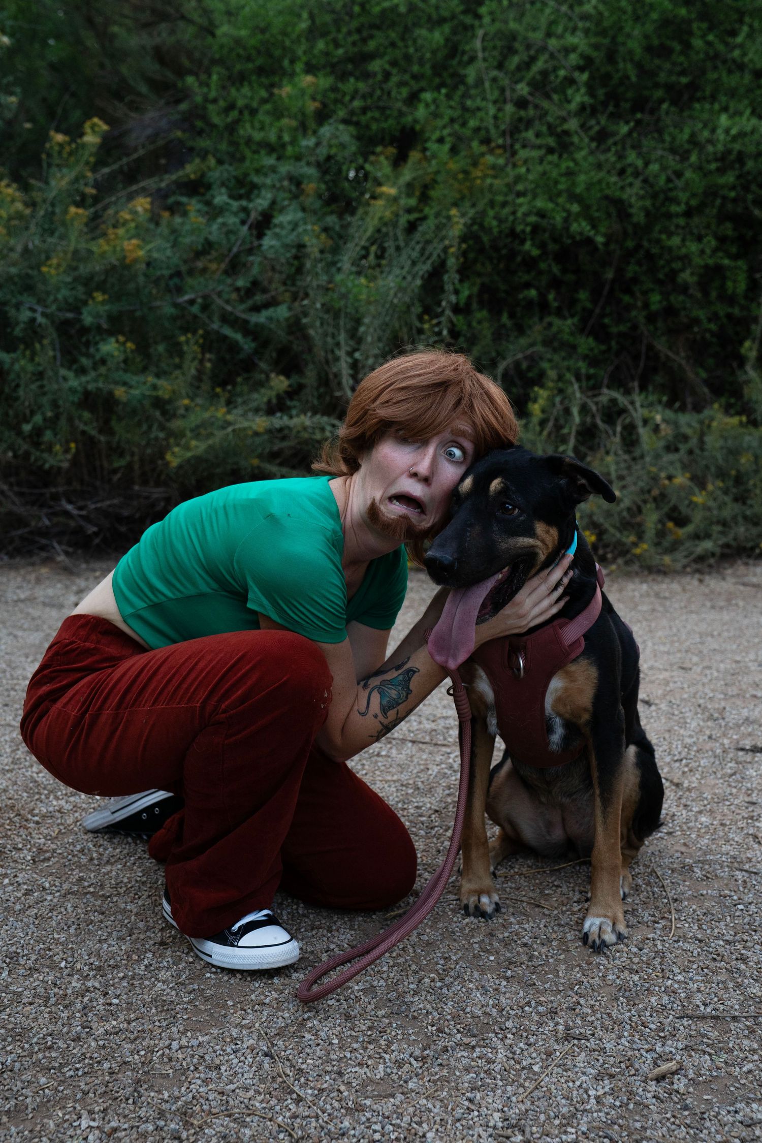 A woman in a green shirt and red corduroy pants crouches on a dirt path, making a funny scared face while hugging a black and tan dog.