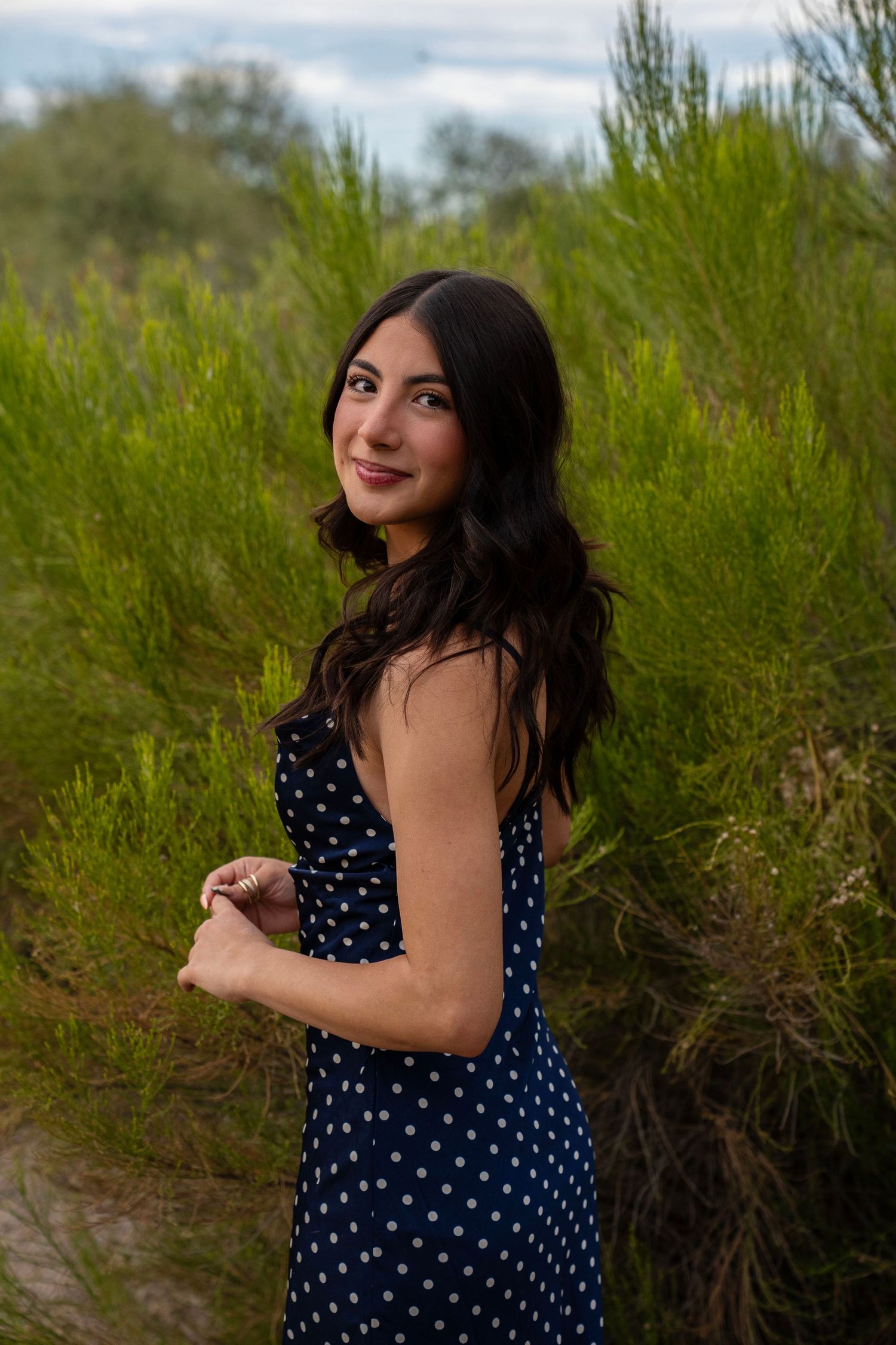 Smiling young woman in navy polka dot dress among green desert shrubs.