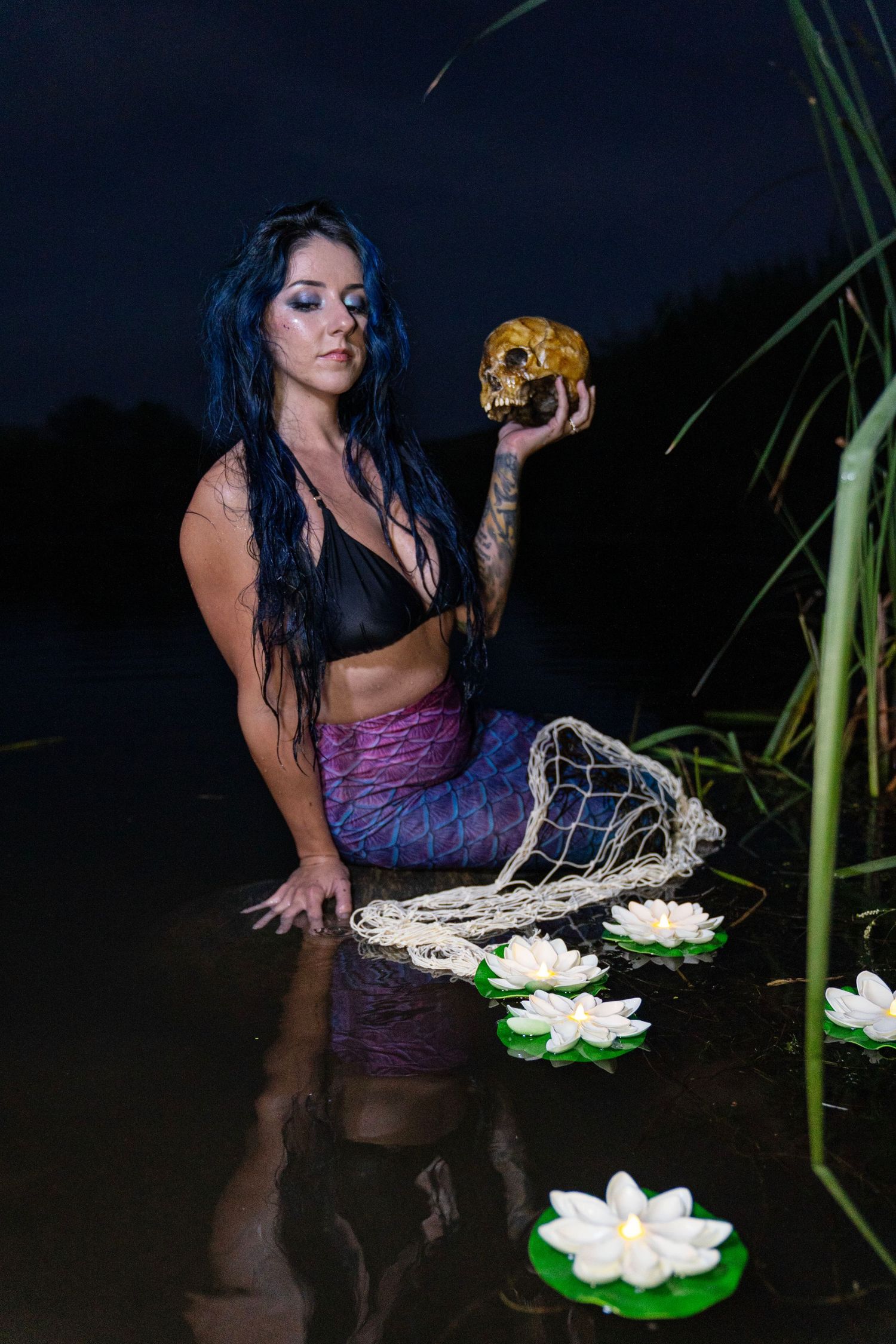 Tattooed woman in mermaid tail holds skull in moonlit water with lotus flowers.