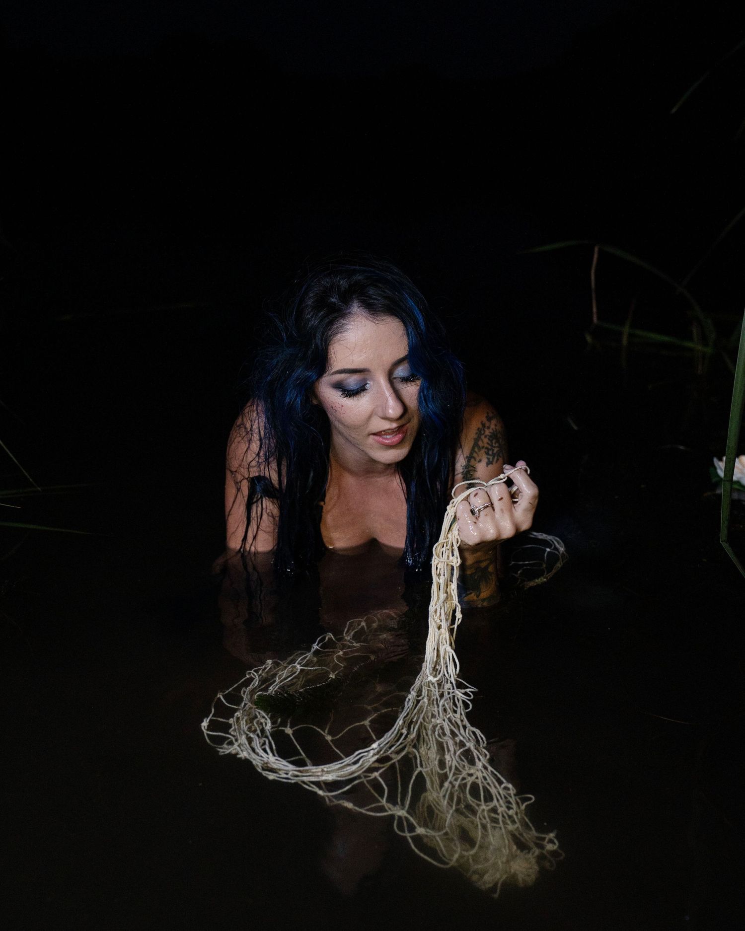 A blue-haired woman in dark water holds a fishing net in a moody nighttime scene.