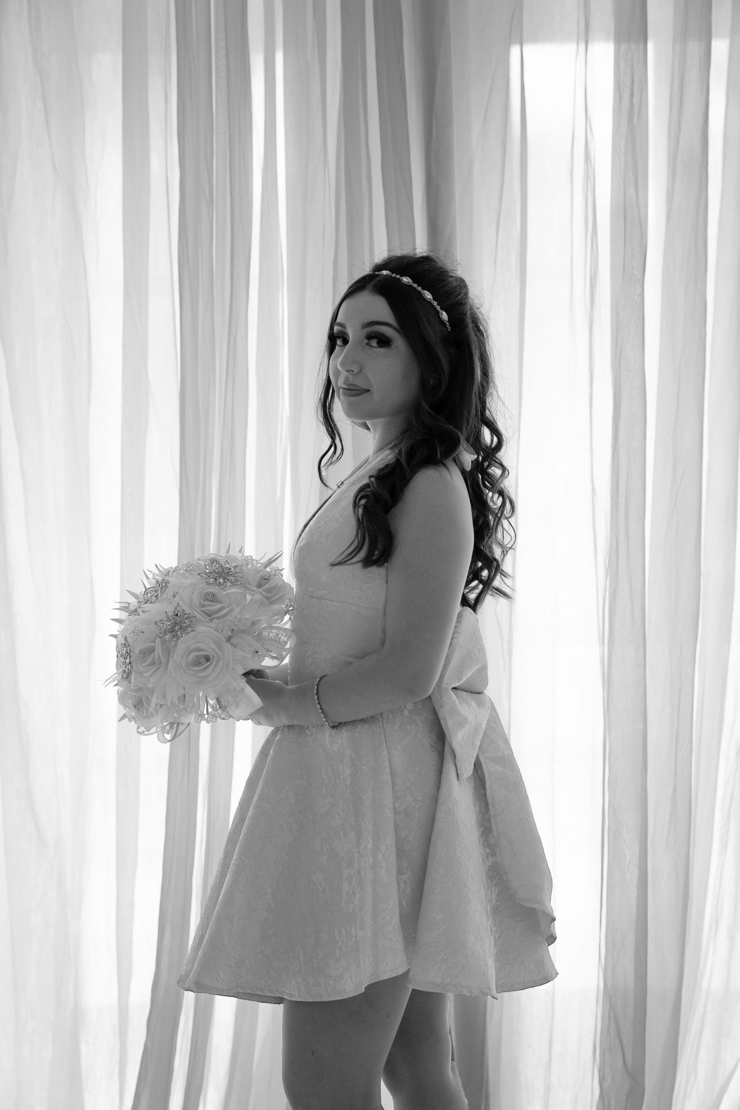Black and white photo of a bride in a short dress holding a bouquet, posing elegantly by sheer white curtains.