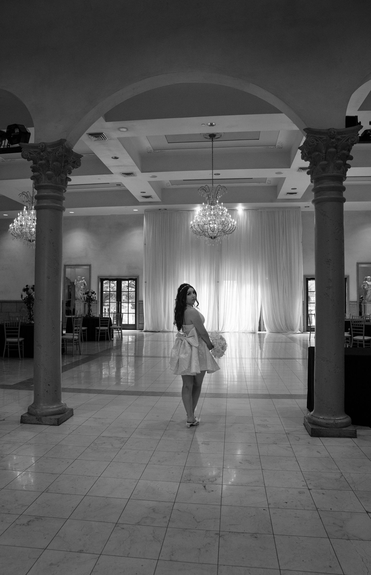 Elegant woman in white dress standing in grand ballroom with chandeliers and columns, black and white photo.