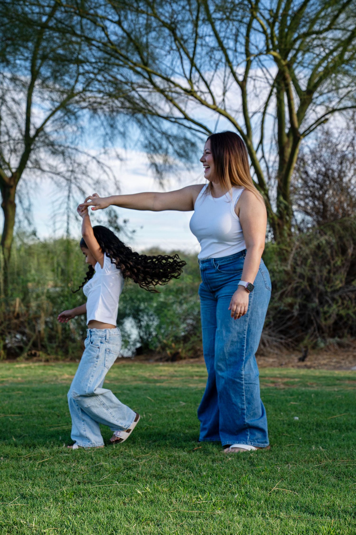 A woman and young girl in matching white tops and jeans dance together on green grass in a park.