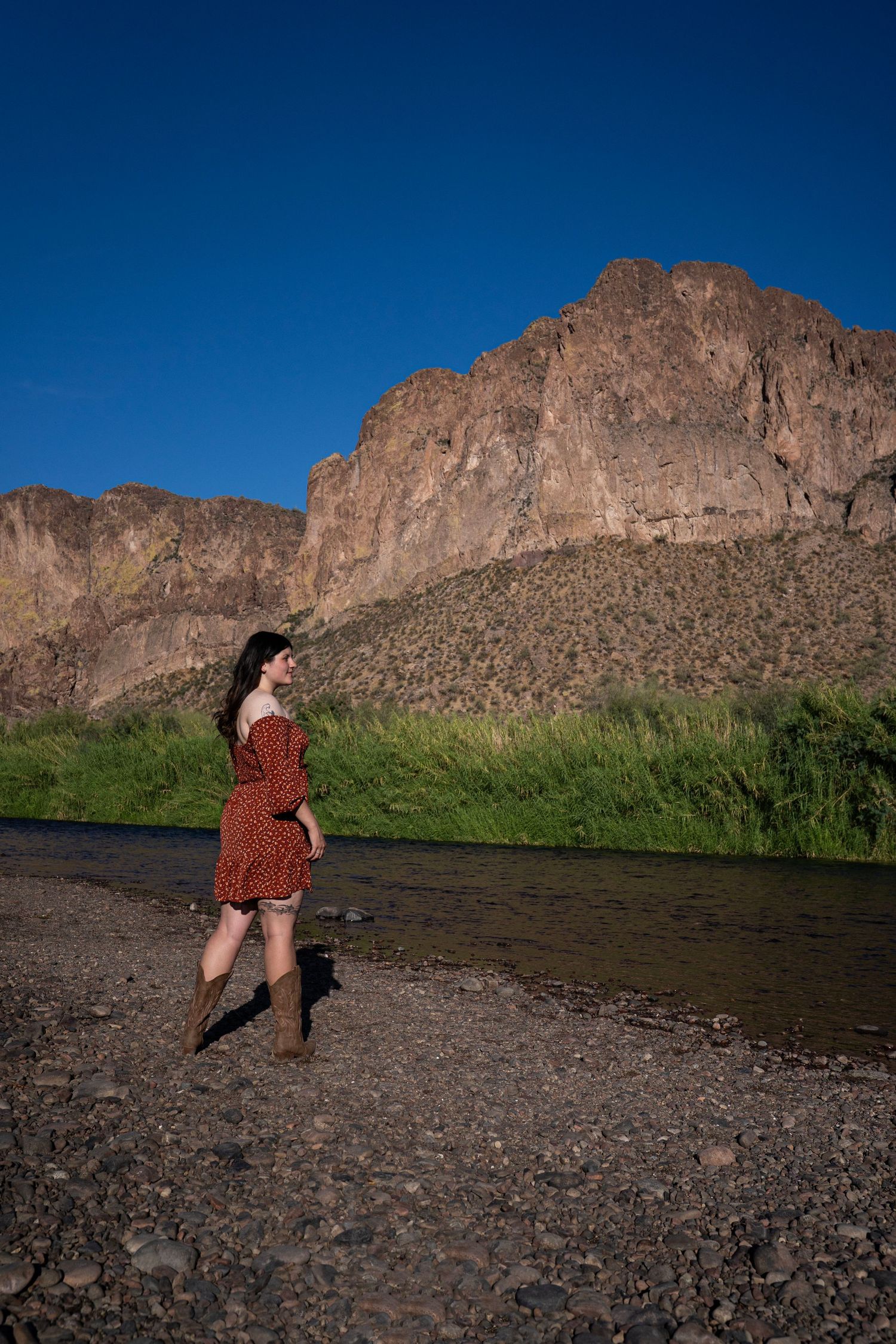Woman in red dress and boots stands by a river with dramatic rocky mountain cliffs under a clear blue sky.