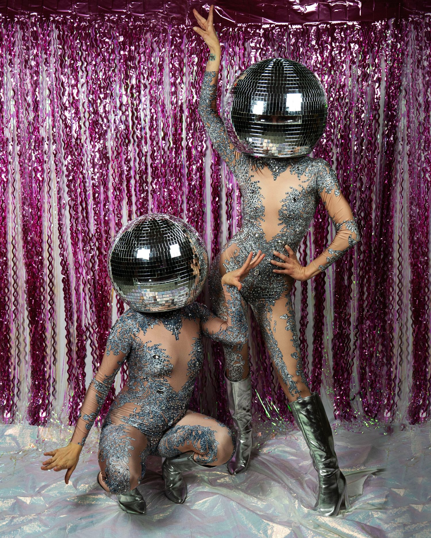 Two performers in glittery bodysuits and disco ball helmets pose against a pink tinsel curtain backdrop.