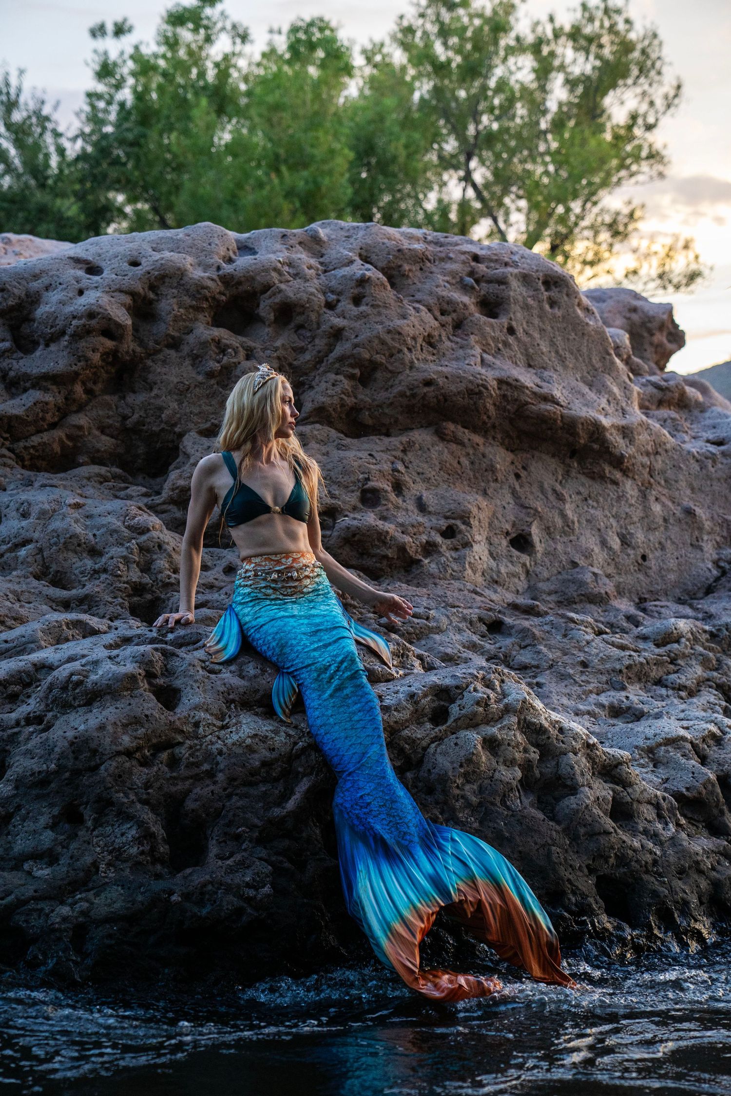 Woman in blue mermaid tail costume reclines on rugged rocks near water at sunset. Mermaid photoshoot, fantasy photoshoot. Arizona photograph