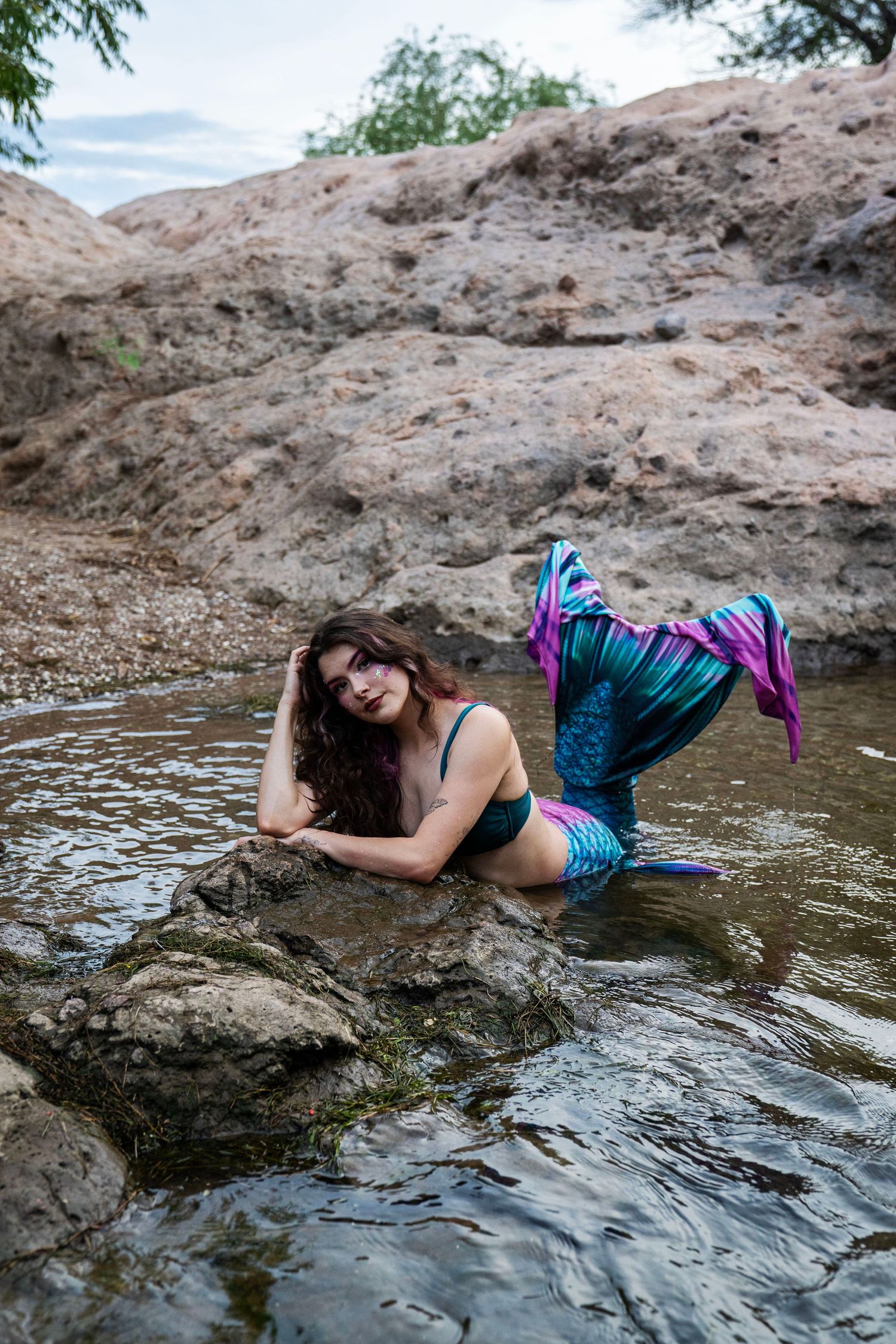 A woman in a colorful mermaid tail costume lies in shallow water leaning on a rock. Fantasy Photoshoot