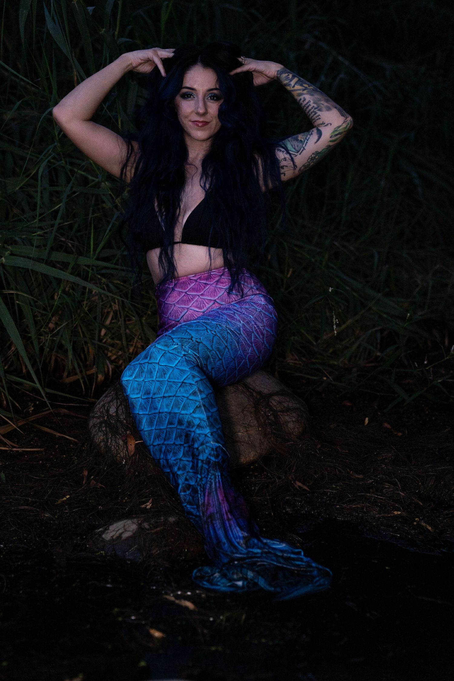 Dark-haired woman in mermaid tail costume poses on rock with hands in hair among green reeds.