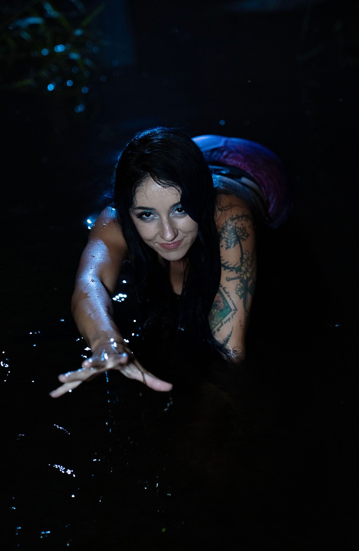 Tattooed woman in mermaid costume splashes in dark water at night reaching toward the camera.