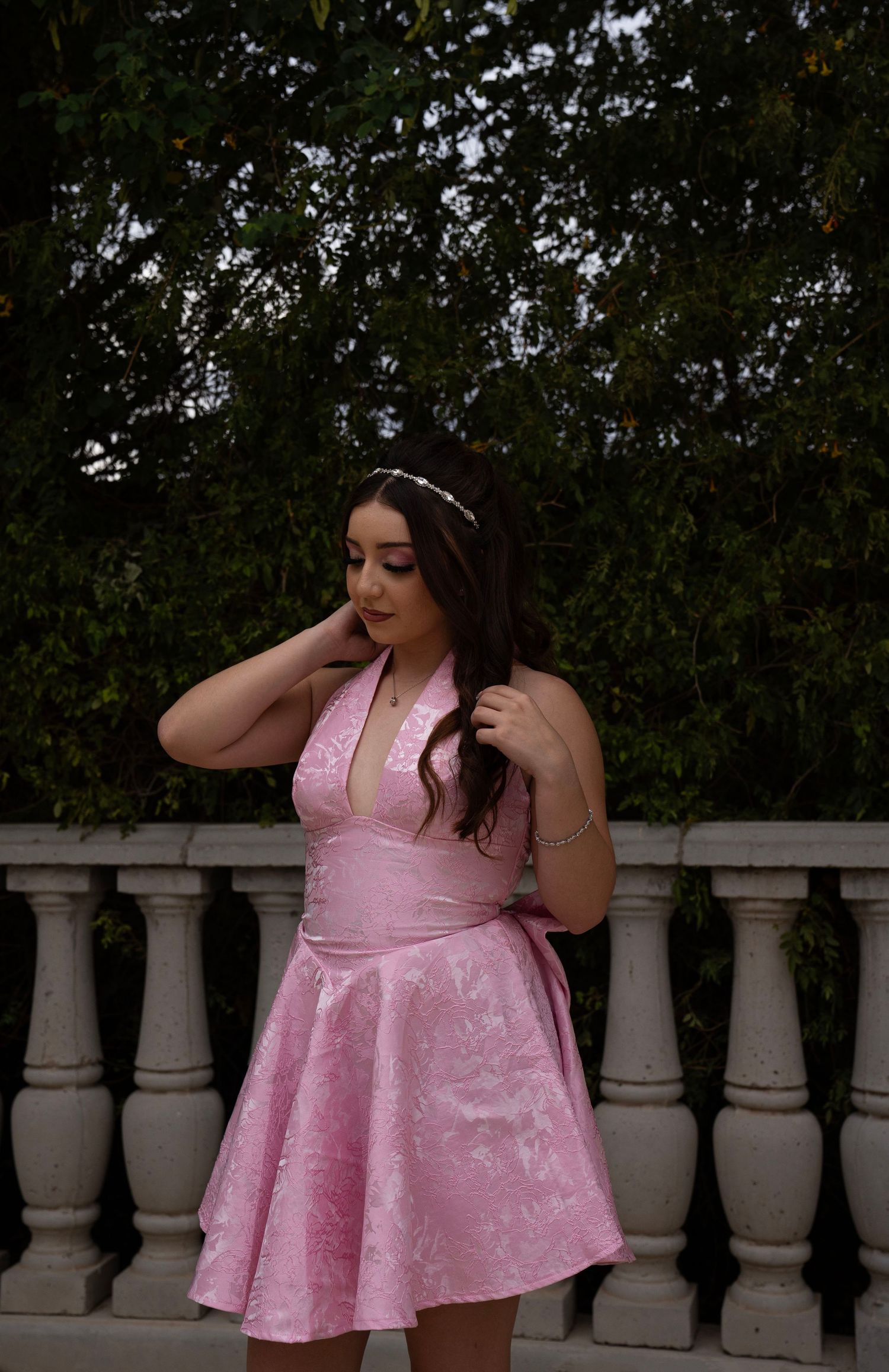 Young woman in pink halter dress standing by white balustrade with dark green foliage background at dusk.