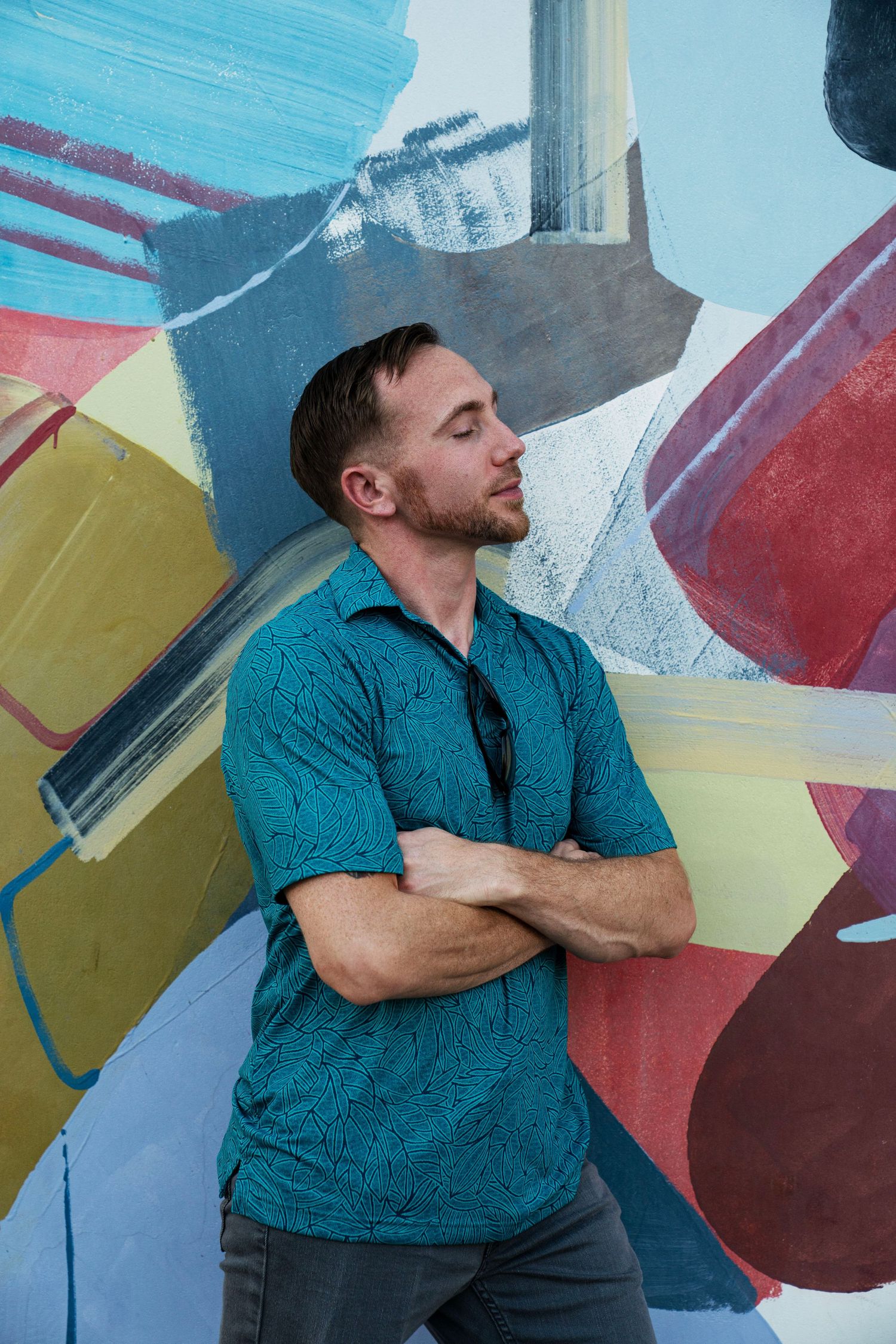 Man in teal shirt with arms crossed leaning against colorful abstract mural wall.