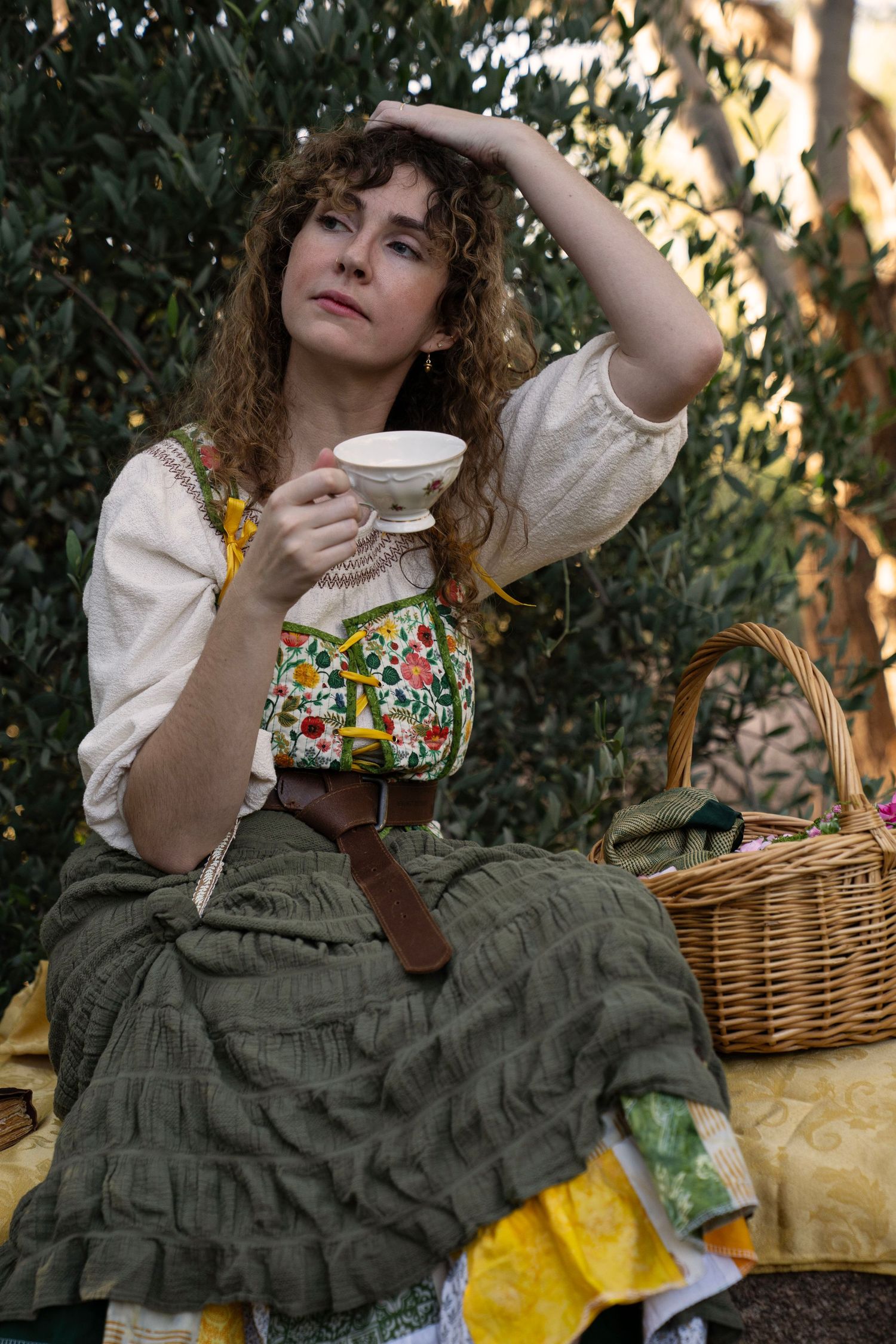 Woman with curly hair in a floral vest and green skirt sits outdoors holding a teacup beside a wicker basket.