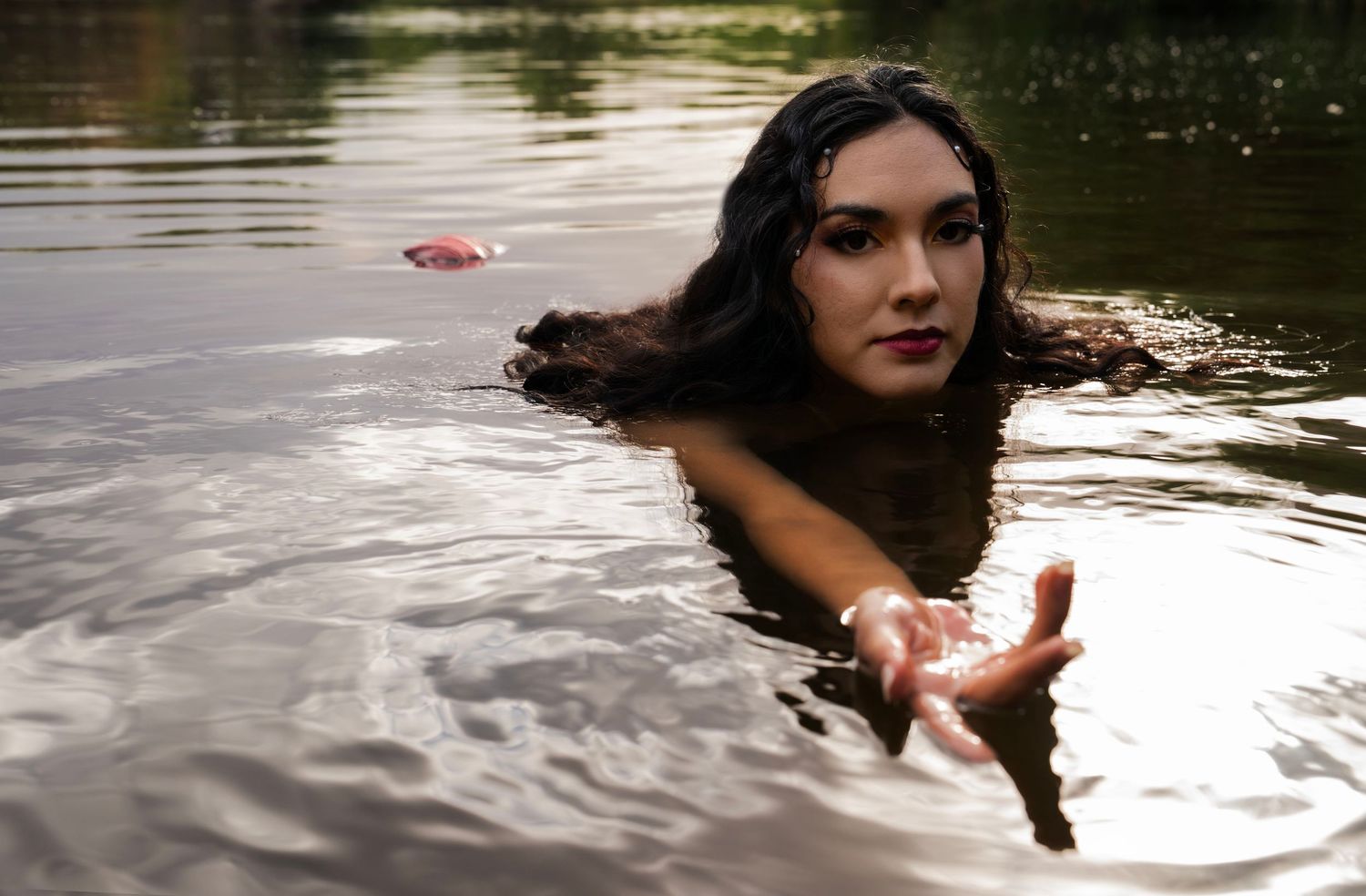 A woman with wet dark hair emerges from a river, reaching her hand toward the camera in a dramatic water photoshoot.