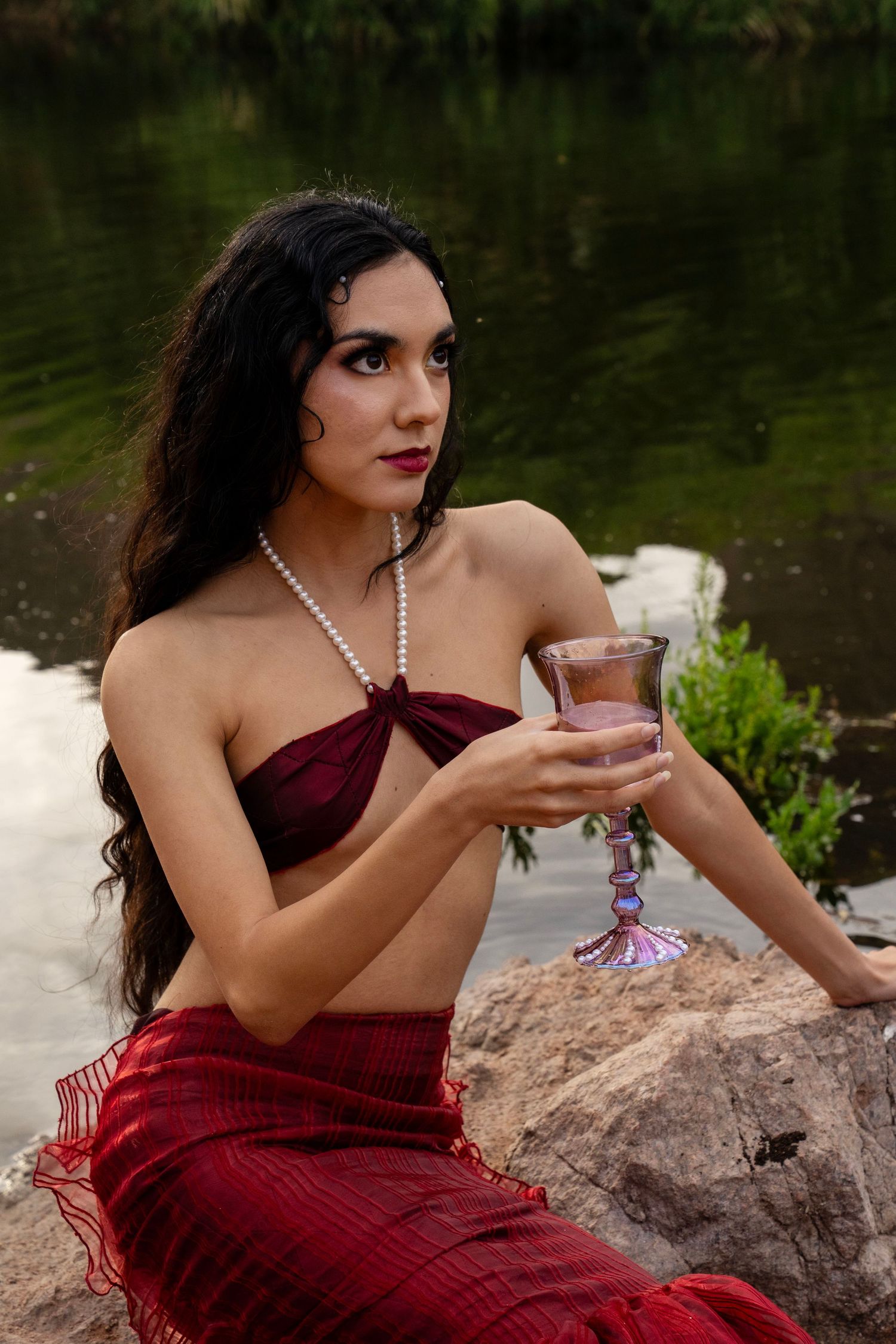 A woman in a red mermaid costume and pearl necklace holds an ornate glass of pink liquid near a river.