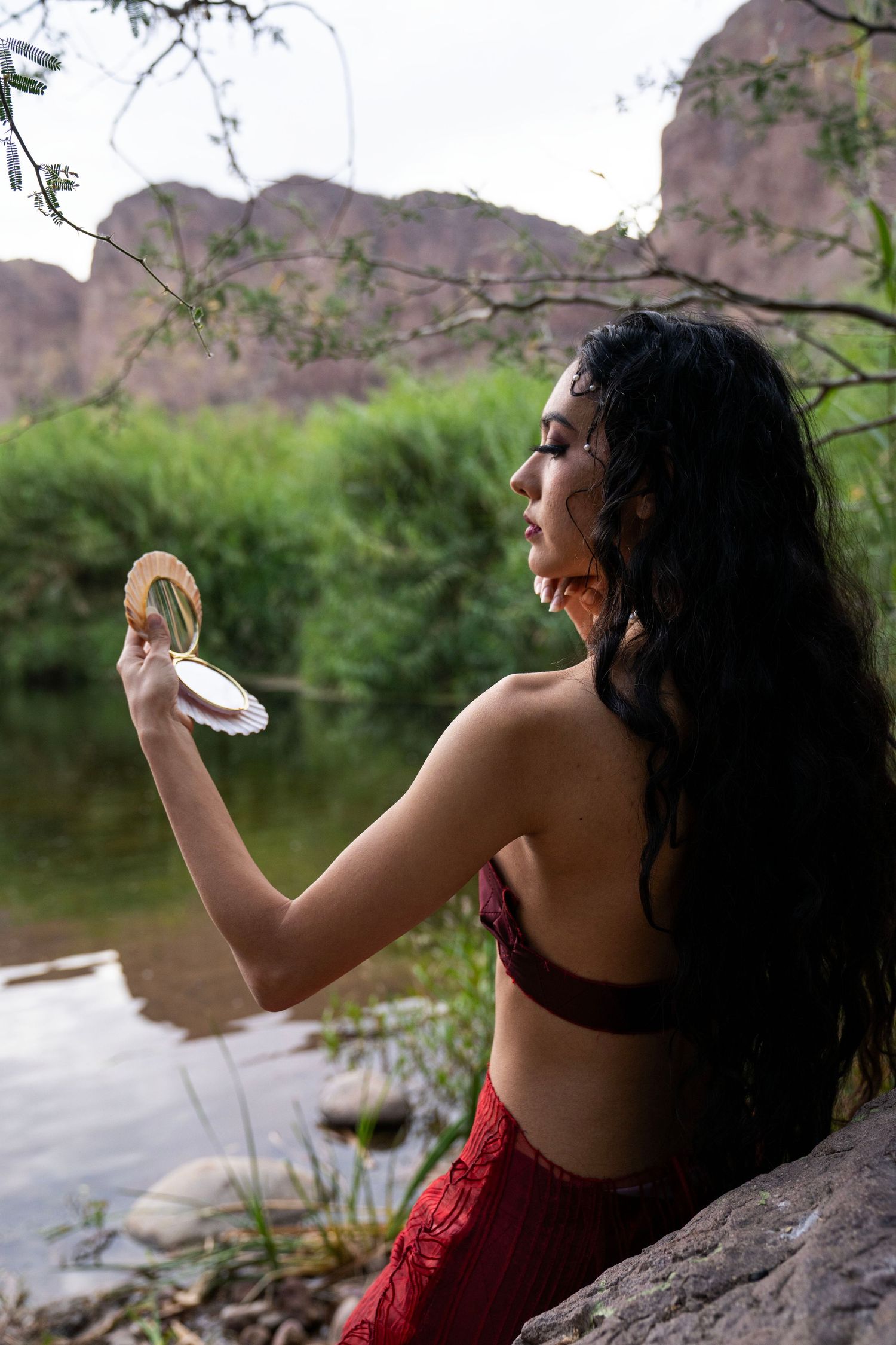 Woman in red mermaid costume holding a seashell mirror while sitting on rocks near a river.