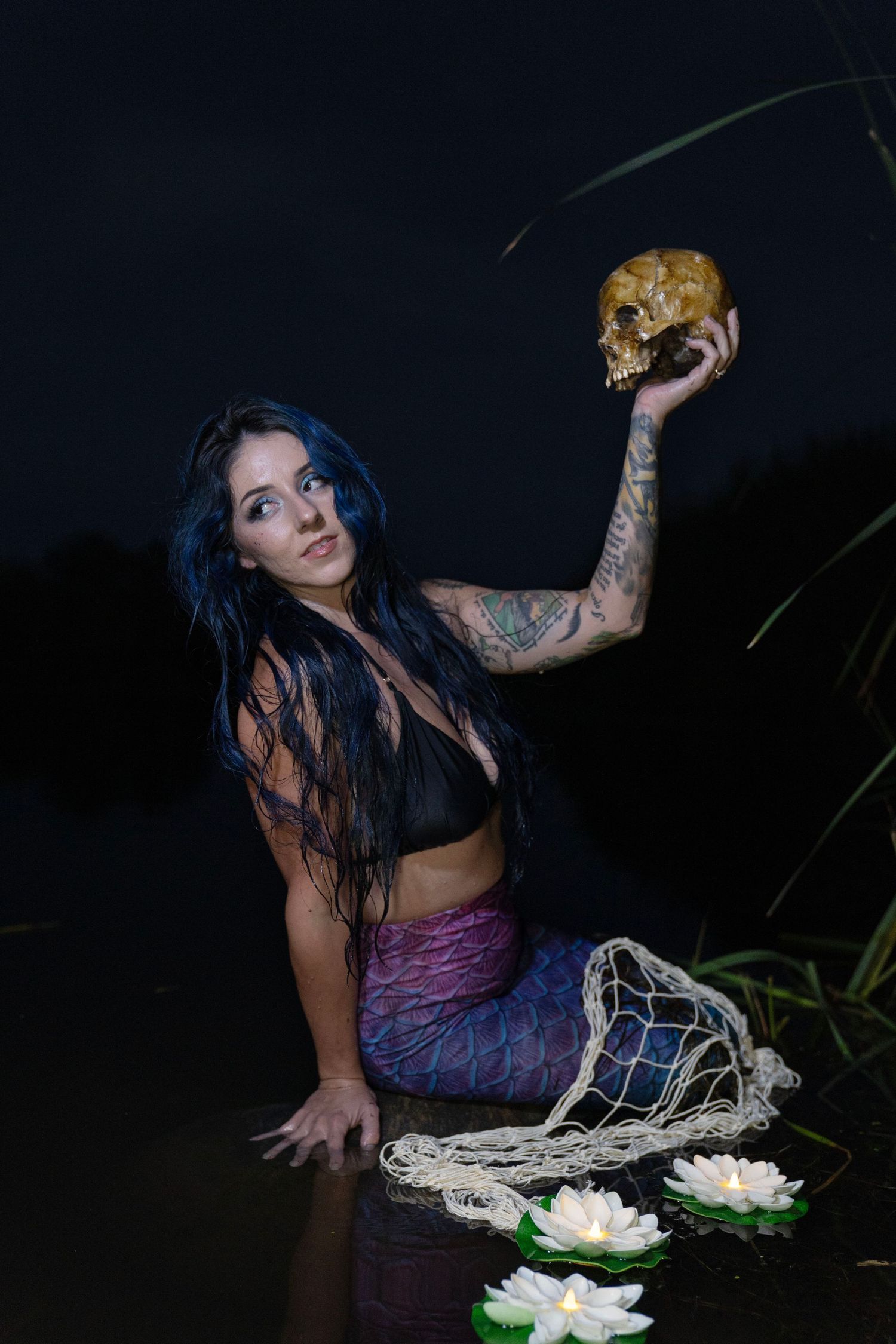 Tattooed woman with blue hair holds a skull while sitting in water at night surrounded by lotus flowers.