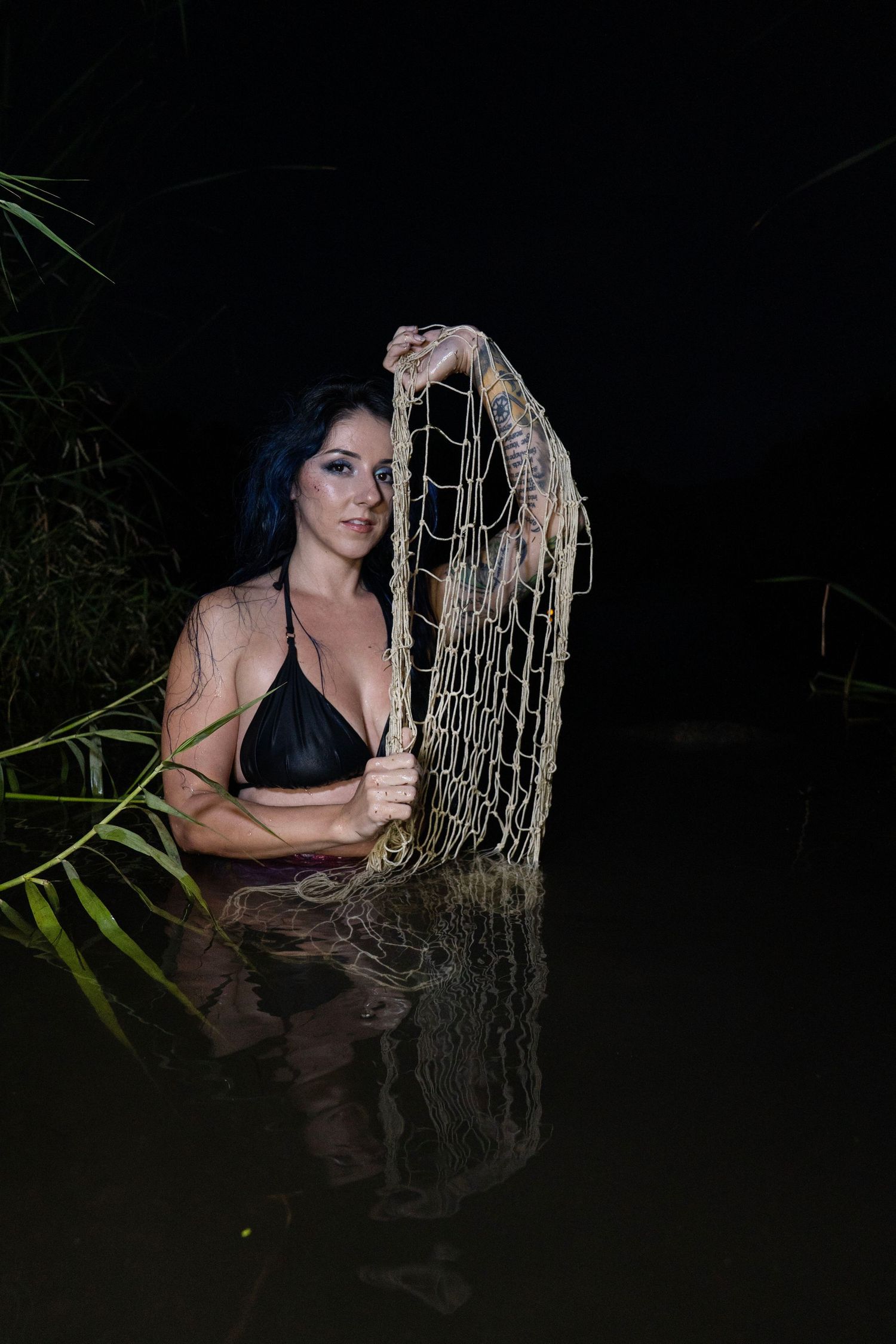 Woman in black bikini holding fishing net while standing in dark water at night.