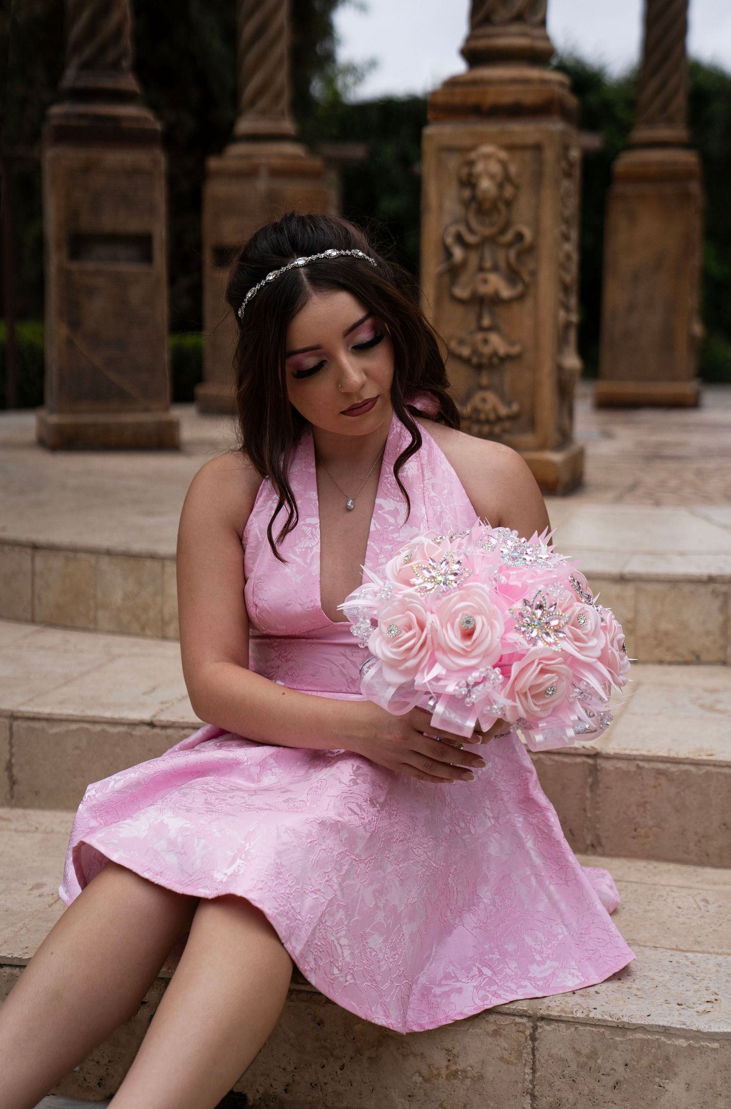 Young woman in pink lace halter dress sitting on stone steps holding a pink floral bouquet with rhinestone brooches.