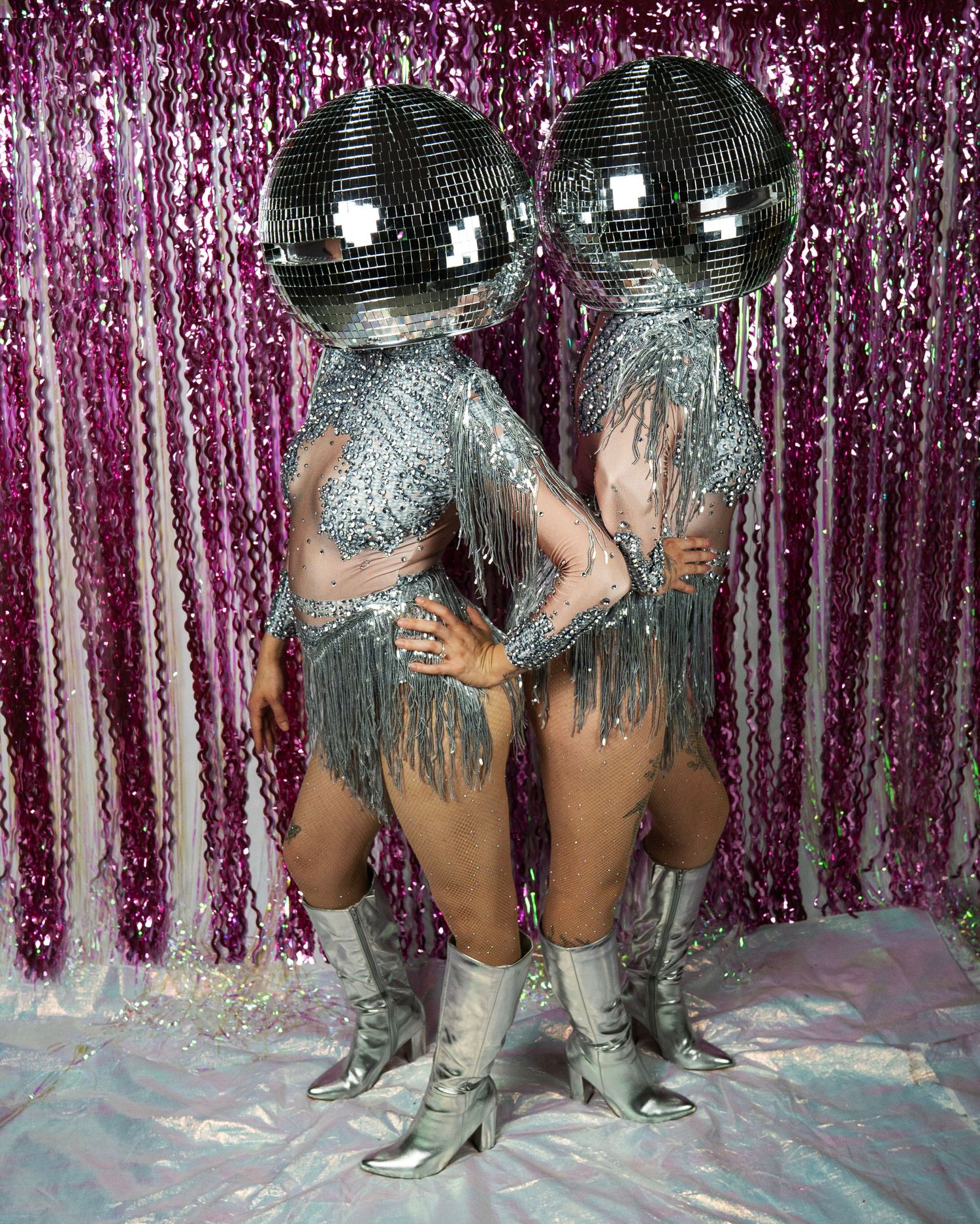 Duo in matching silver crystal fringe outfits with disco ball heads embrace in silver ankle boots.