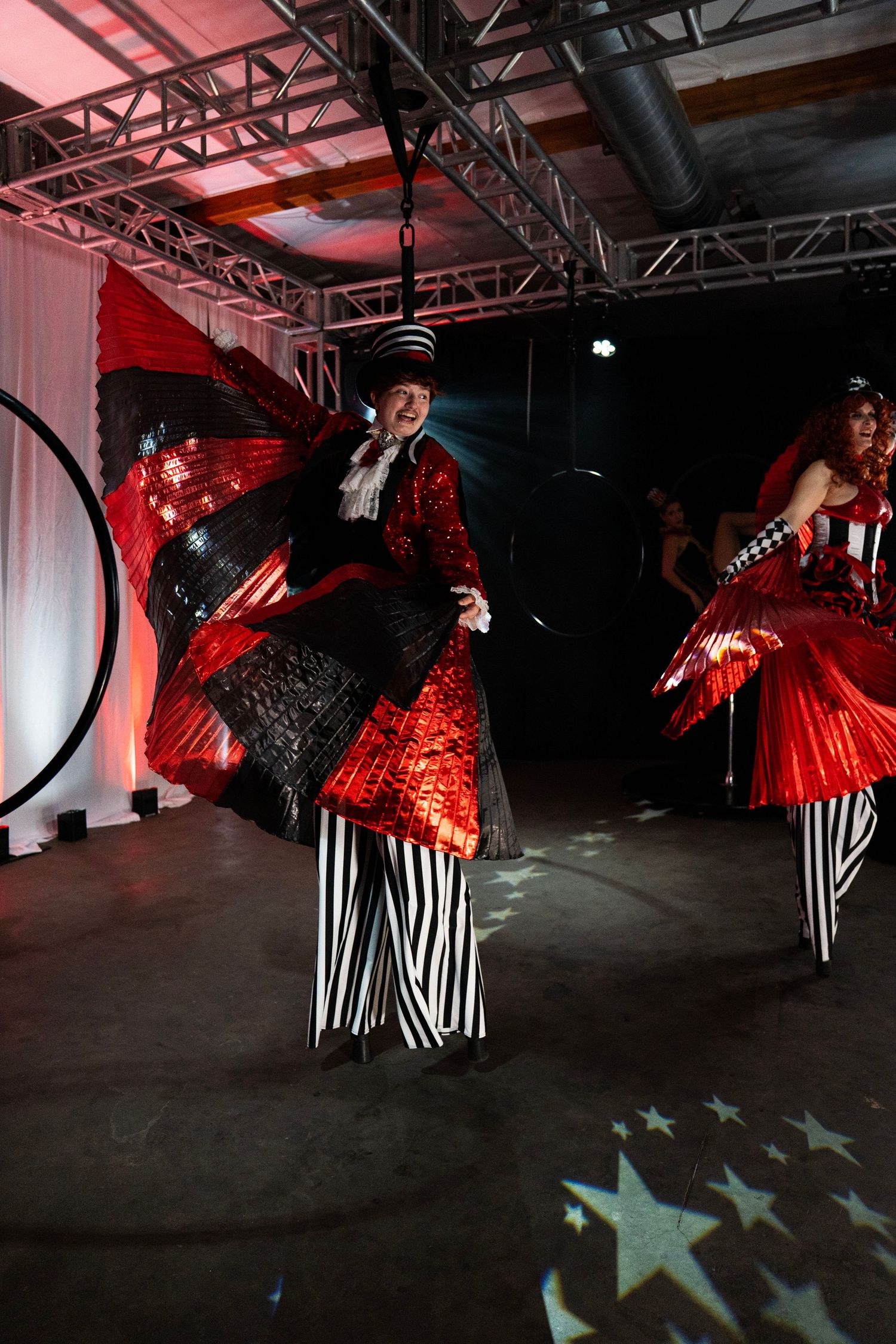 Two circus stilt walkers in red sequin costumes perform with butterfly wings at event.