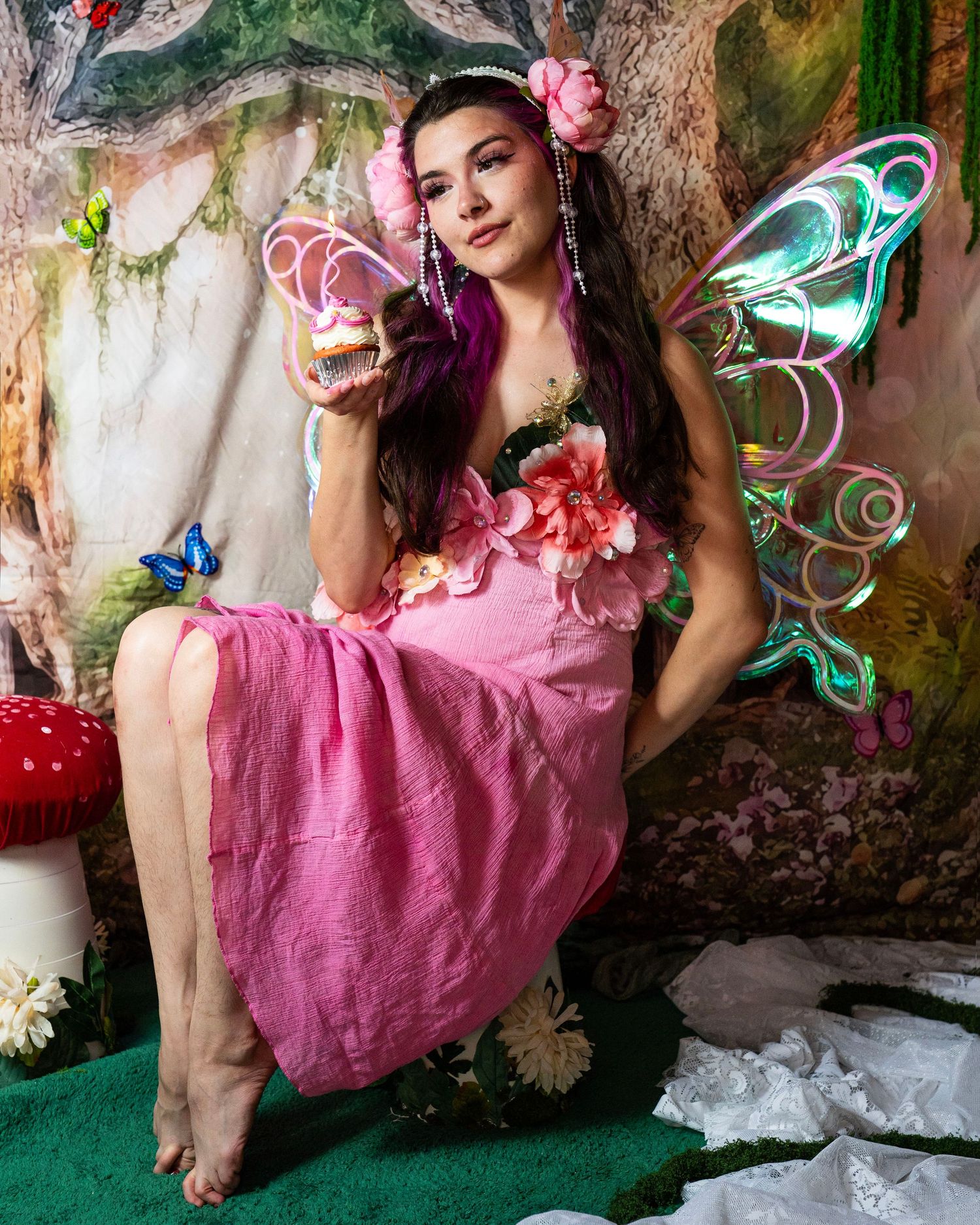 Fairy costume model in pink floral dress with iridescent wings holds cupcake in magical forest backdrop. Fantasy Photoshoot
