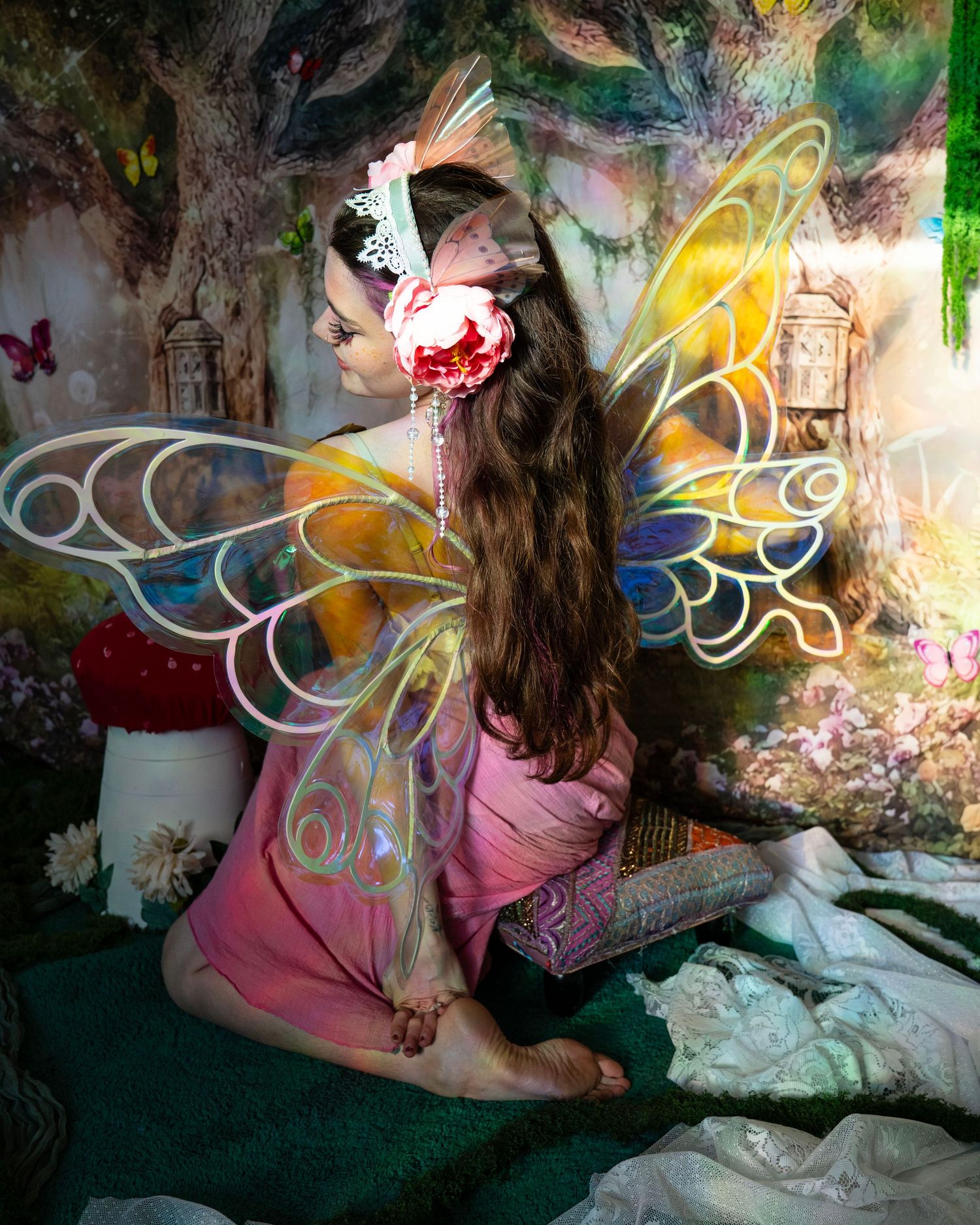 Woman in pink dress with iridescent fairy wings and floral hair accessory, seated in magical forest photo studio. Fantasy Photoshoot