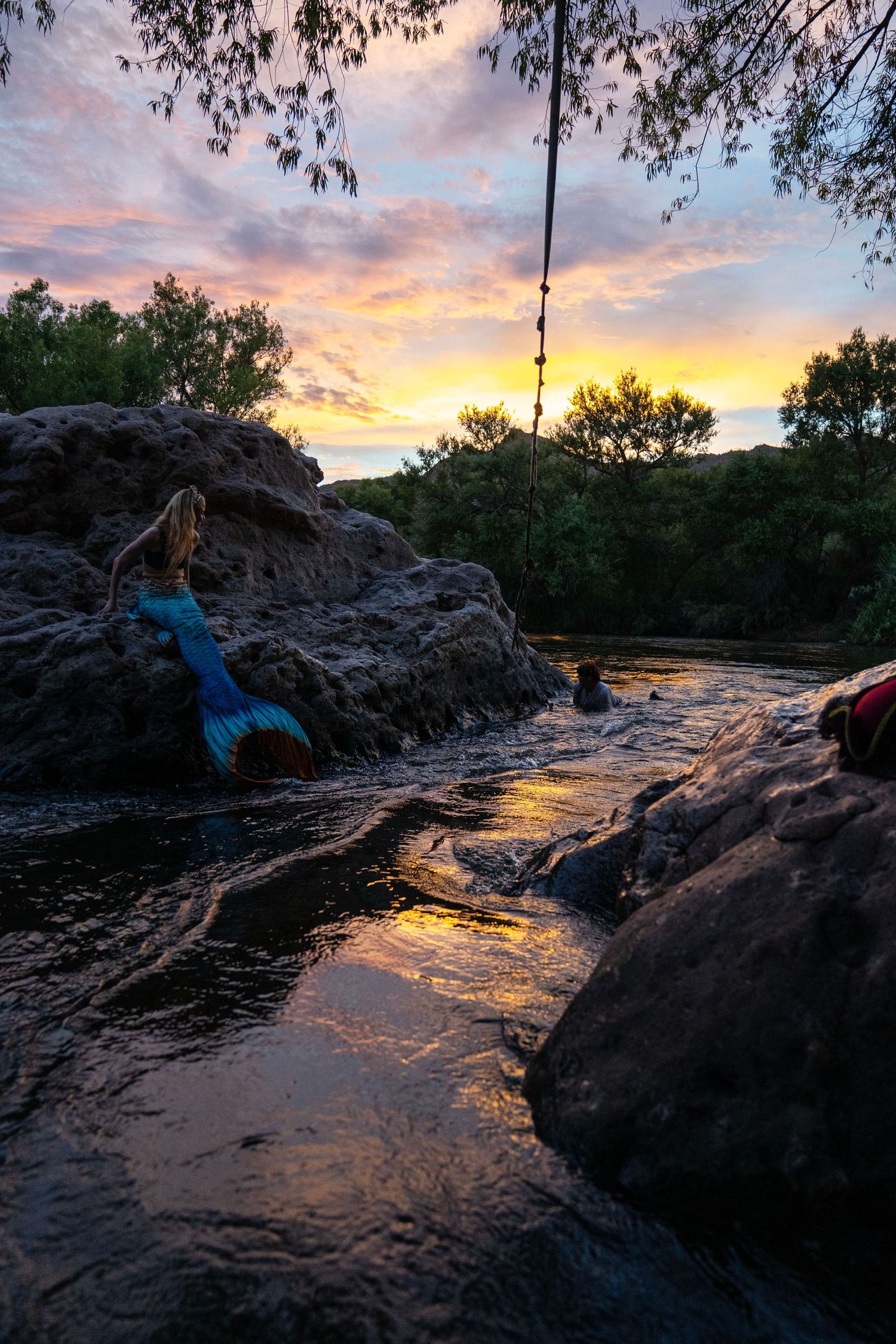 Mermaid costume figure resting on rocks beside flowing river at golden sunset. Mermaid photoshoot, fantasy photoshoot. Arizona photographer.