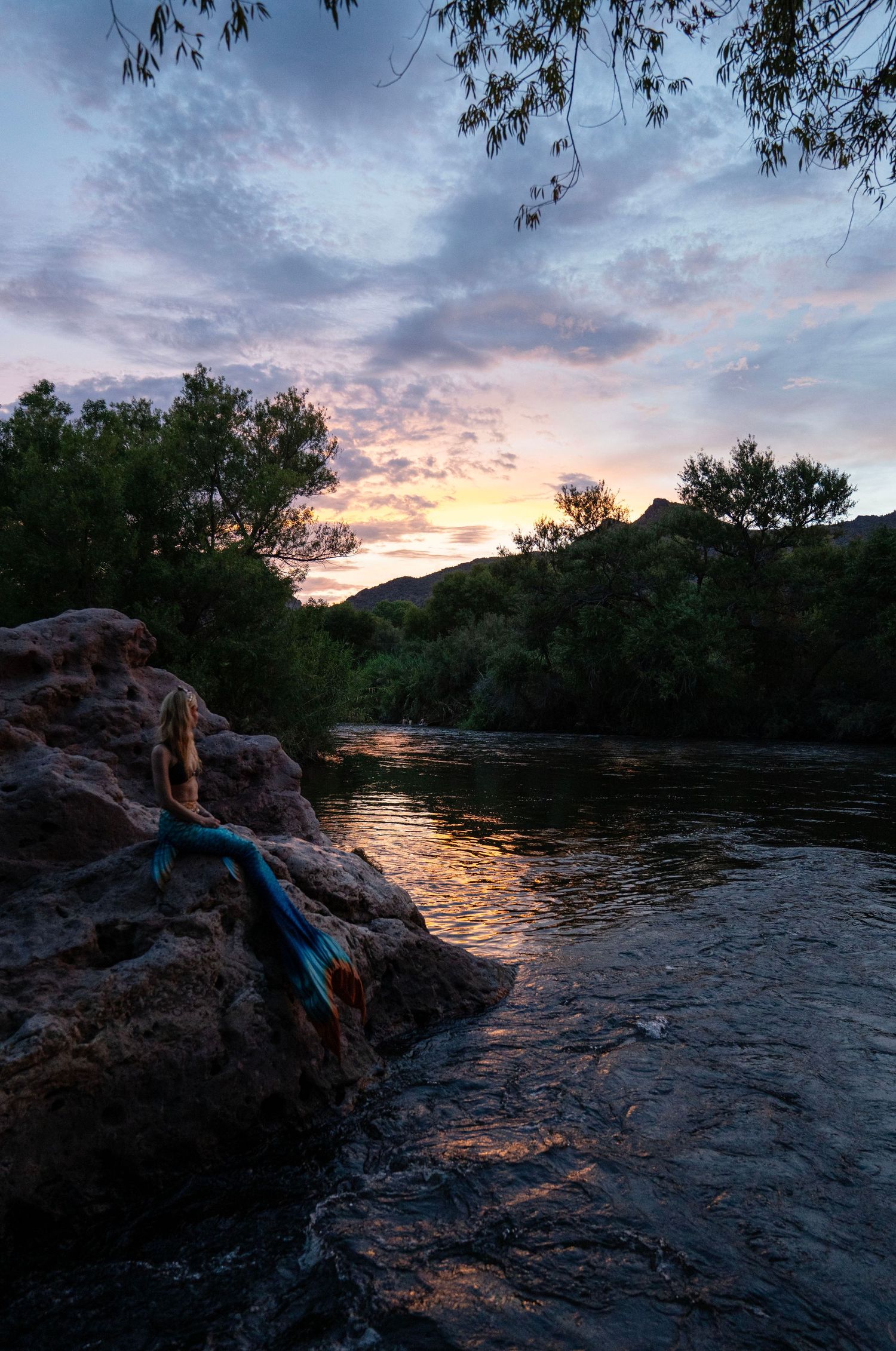 Person in blue mermaid tail sitting on rocks by a river under dramatic sunset sky.Mermaid photoshoot, fantasy photoshoot. Arizona photograph