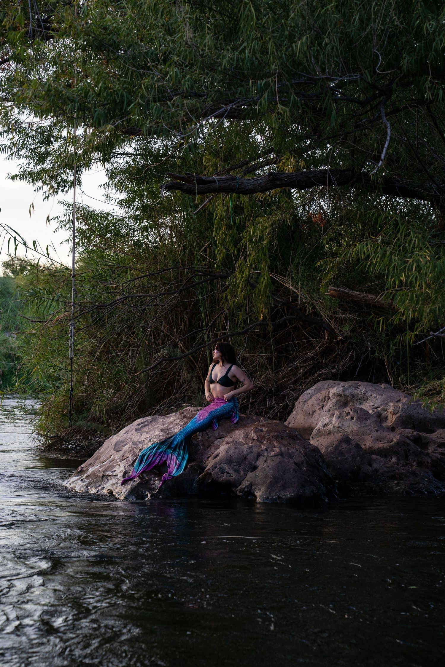 A woman in a mermaid costume with colorful tail sits on rocks by a river surrounded by lush green trees. Mermaid photoshoot, fantasy photosh