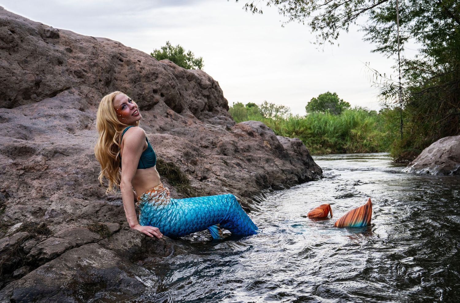 A blonde woman in a blue mermaid tail costume reclines on rocks beside a shallow river. Fantasy Photoshoot