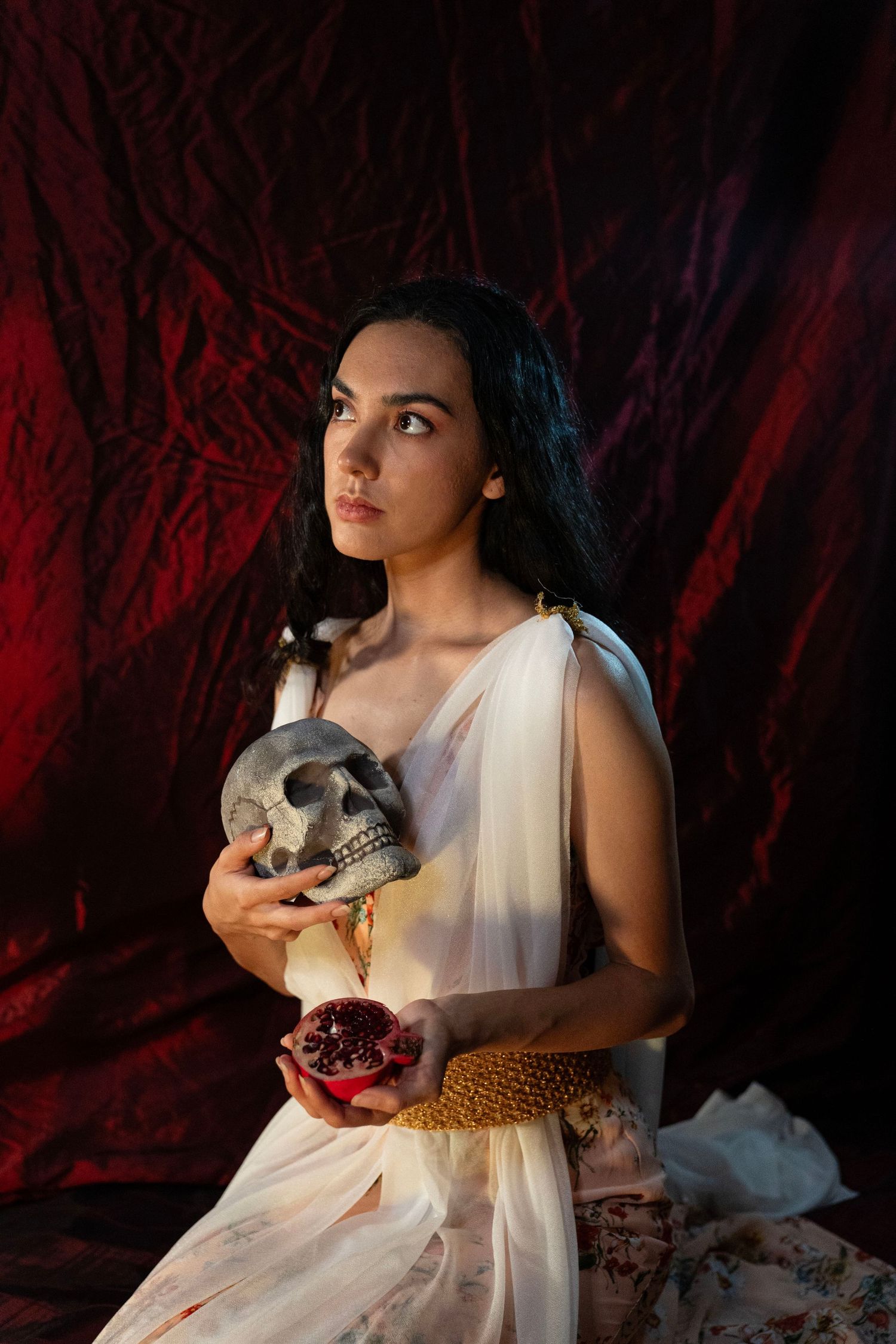 Woman in a flowing white draped gown holds a human skull and a halved pomegranate against a dramatic deep red fabric backdrop.