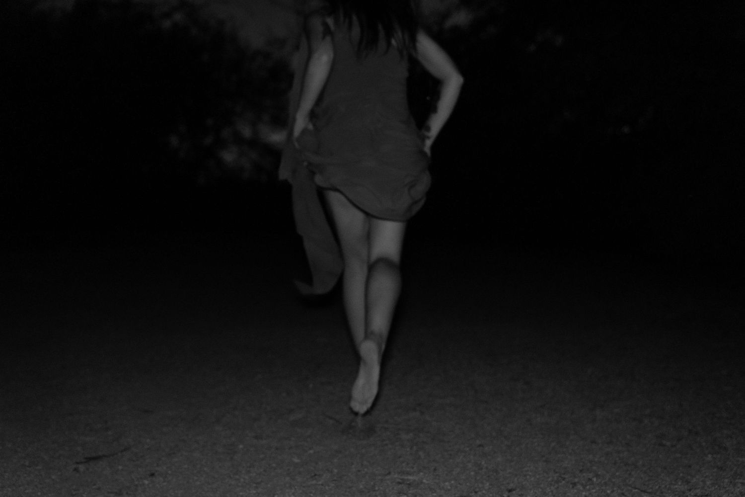 Barefoot woman in a dark dress running away from the camera along a shadowy path at night.