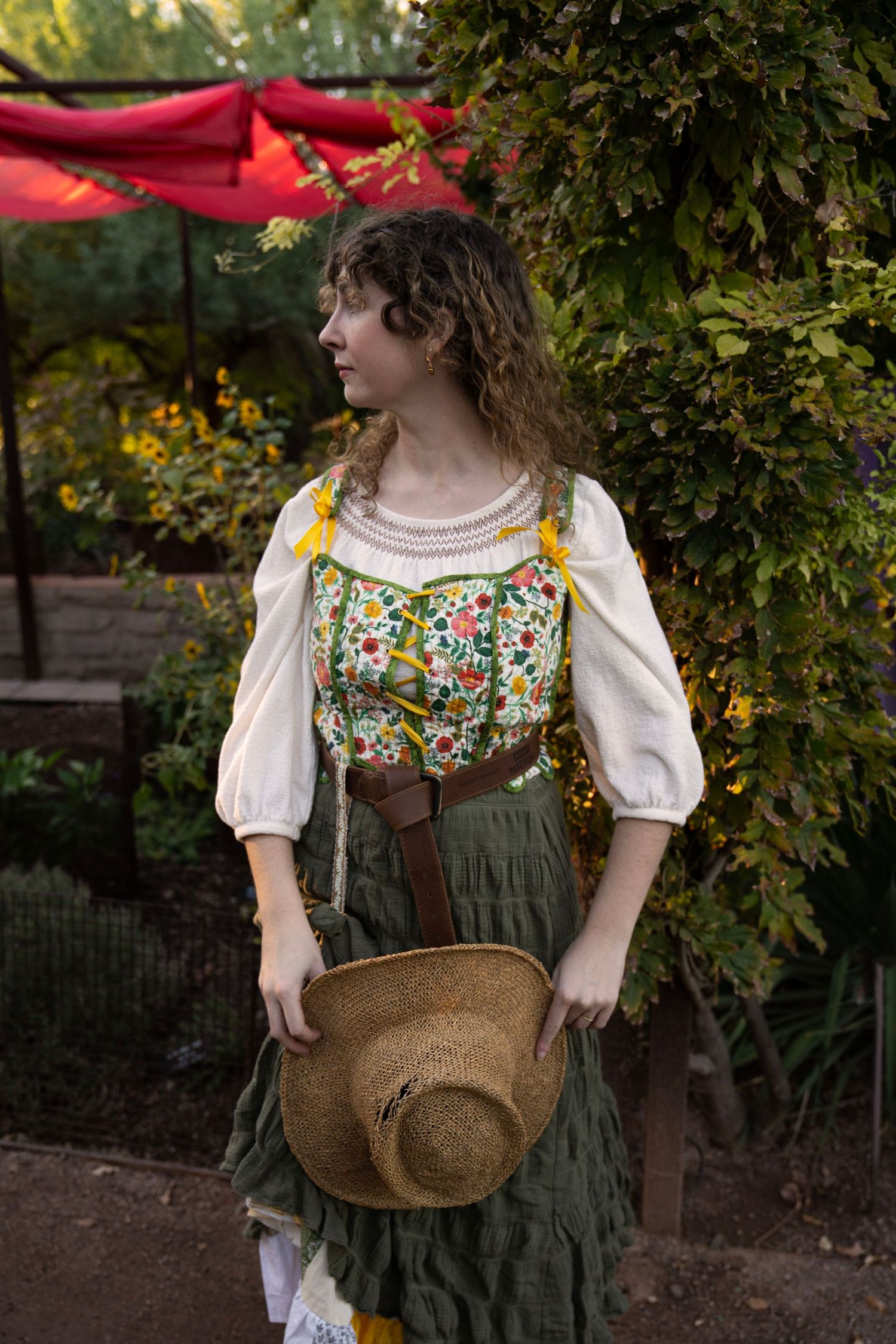 Woman in a white peasant blouse, floral embroidered bodice and green tiered skirt holds a straw hat in a sunlit garden.