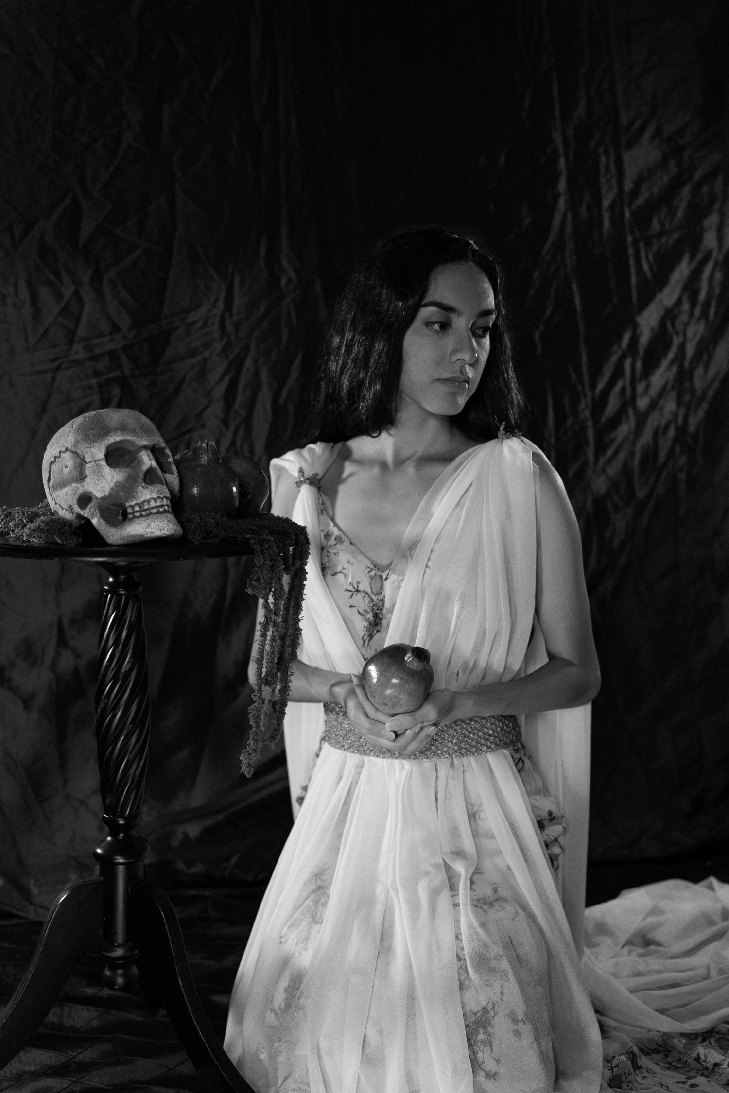 Black and white portrait of a woman in a flowing white gown holding an apple beside a skull on a draped table.