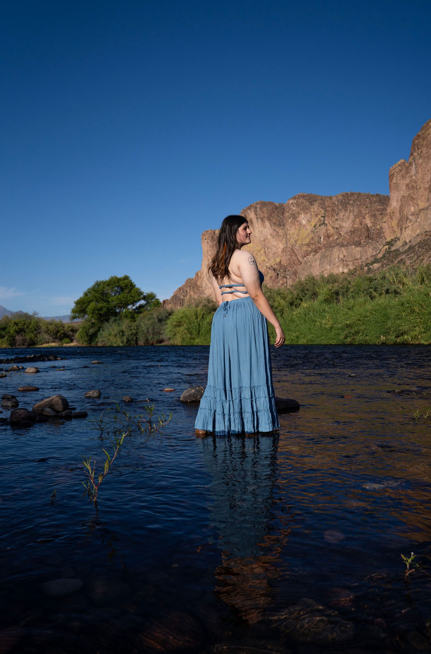 Woman in blue maxi skirt standing in a river with rocky mountains and clear blue sky backdrop.