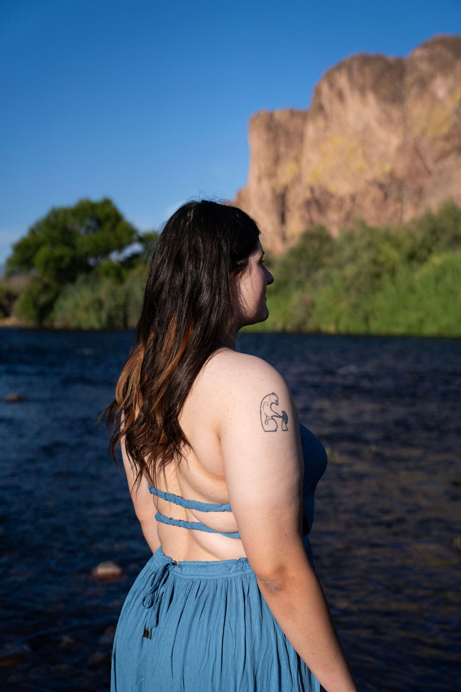 Woman in blue backless dress with tattoo stands by a river with rocky cliffs and green trees under blue sky.