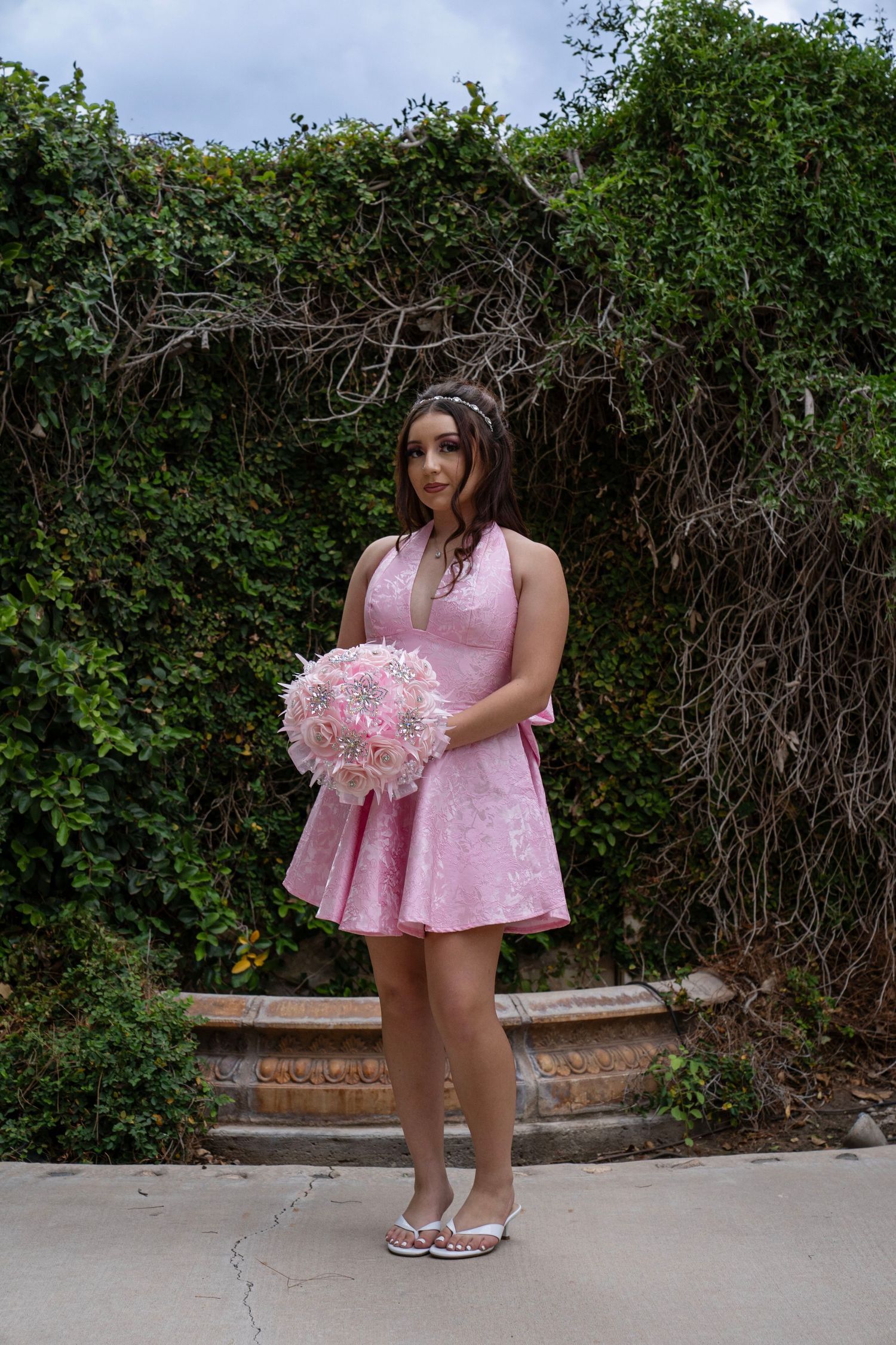 Woman in pink lace dress holding a pink bouquet standing in front of a lush ivy-covered garden wall.