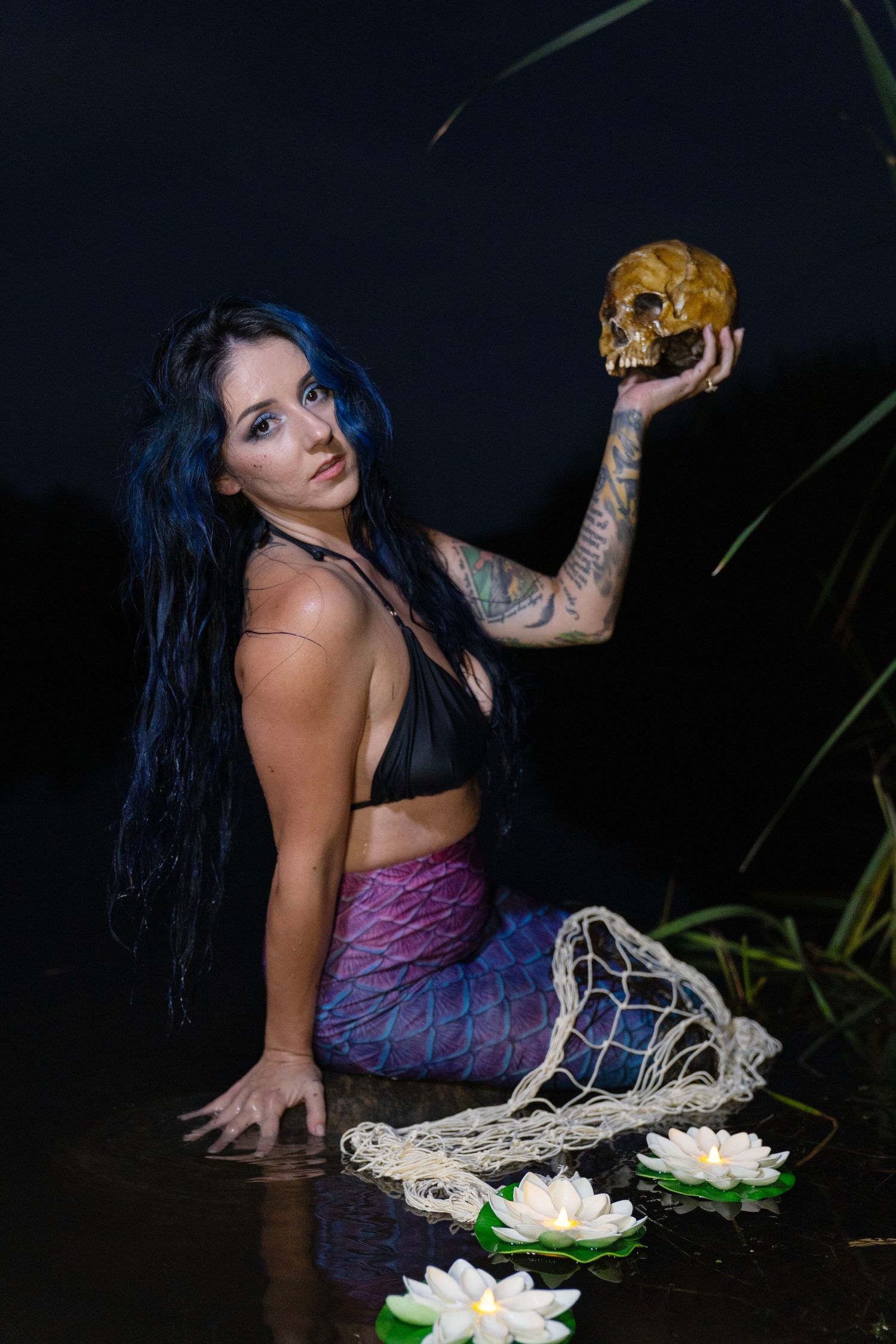 Woman dressed as mermaid holding skull prop with lotus flowers in dark water at night.
