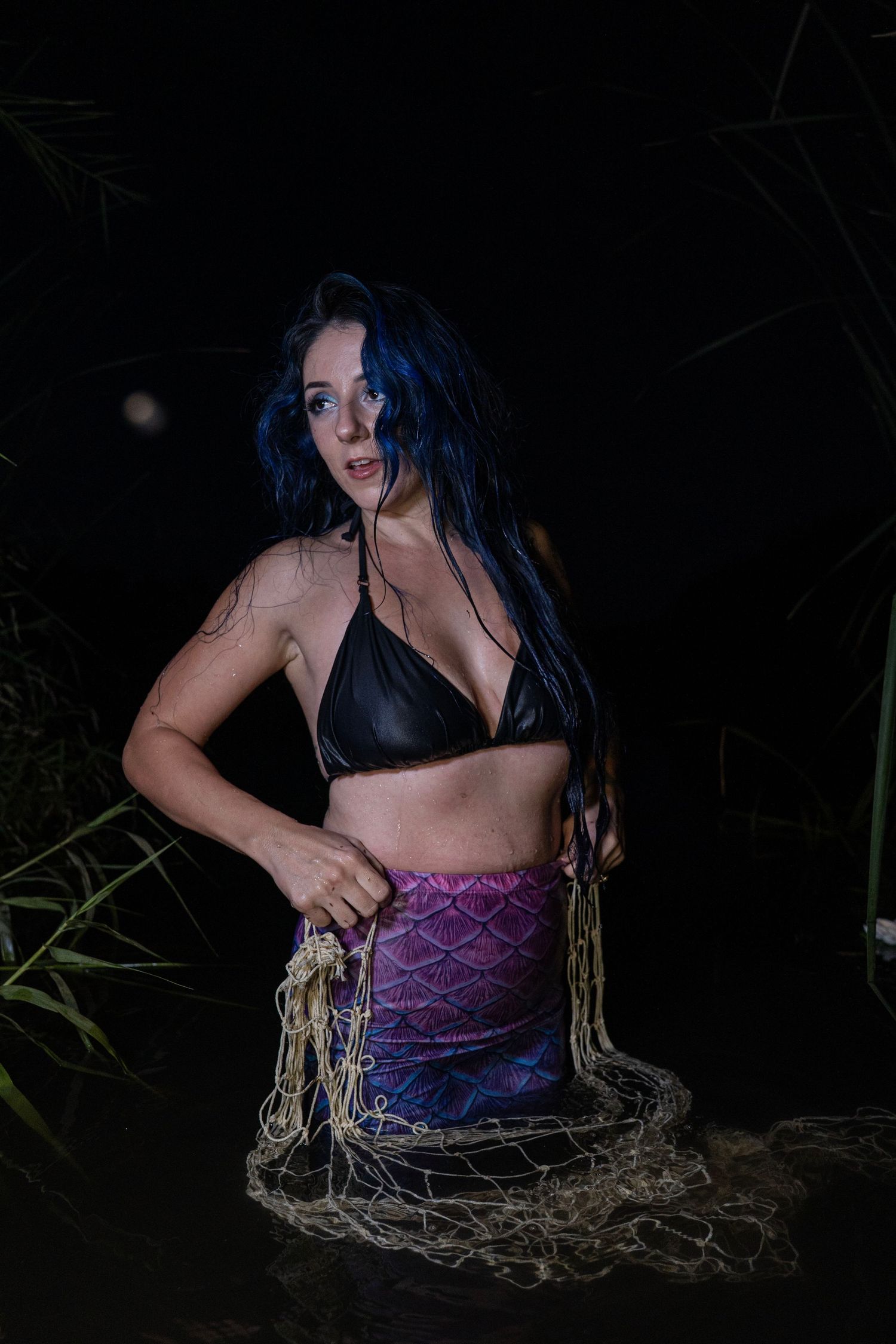 Tattooed woman in black bikini top and mermaid tail holding fishing net in dark water at night.