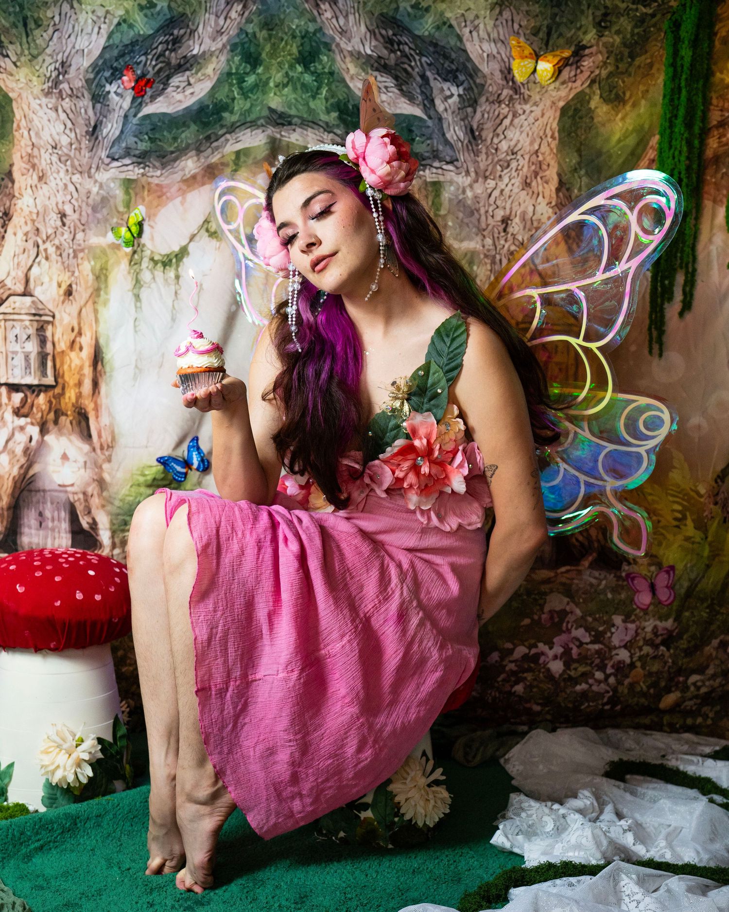 Fairy model in pink floral dress with glowing wings holds cupcake with eyes closed in forest studio. Fantasy Photoshoot