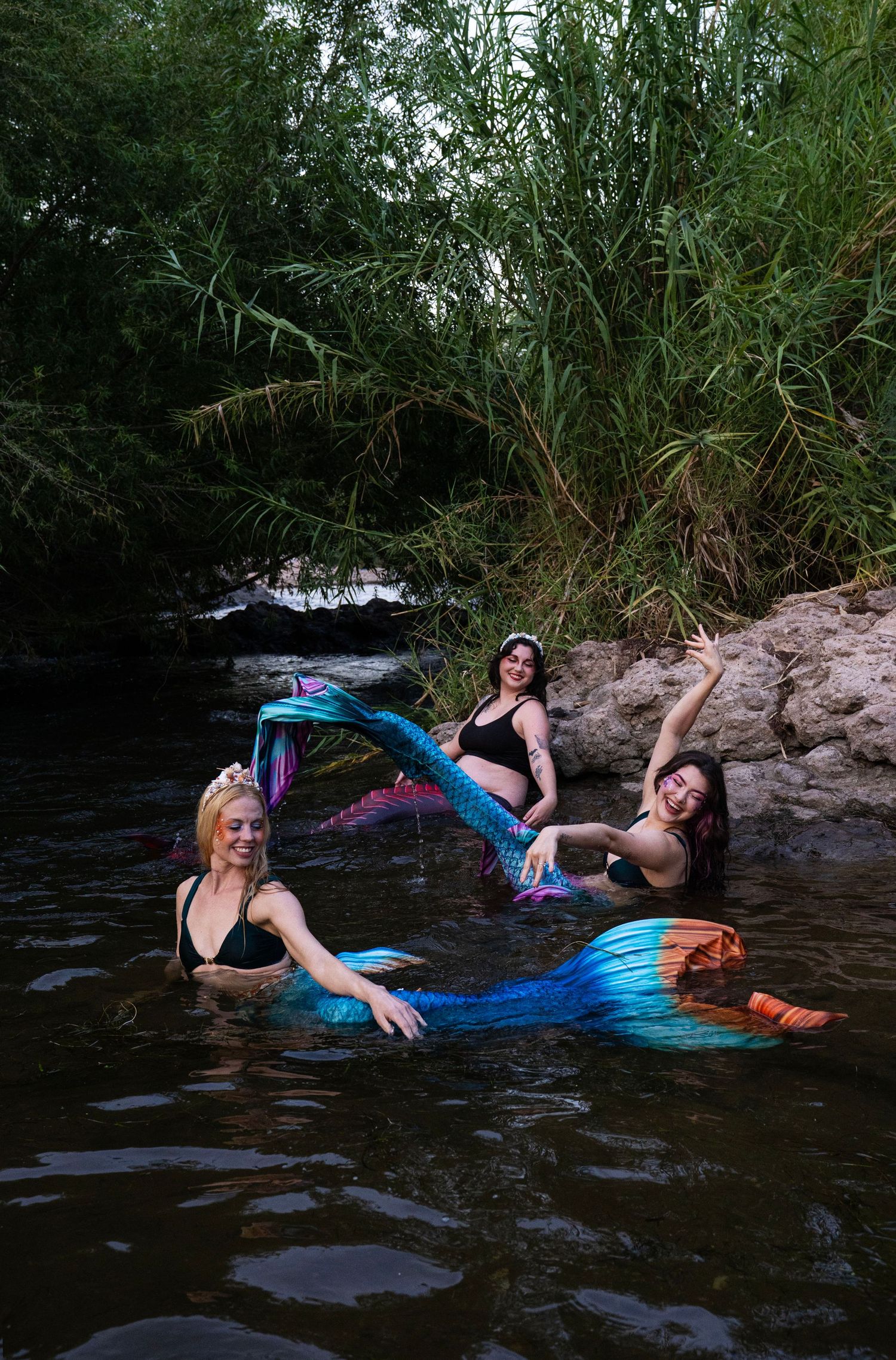 Three women in mermaid tails pose in a natural river surrounded by greenery. Fantasy Photoshoot