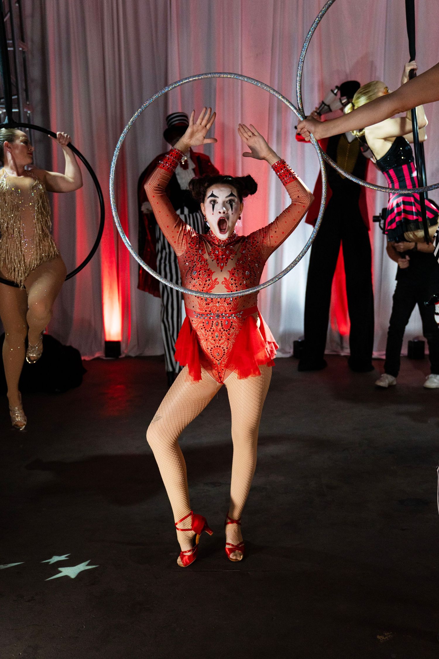 A circus clown performer in red sequined costume poses with a large silver hula hoop on stage.
